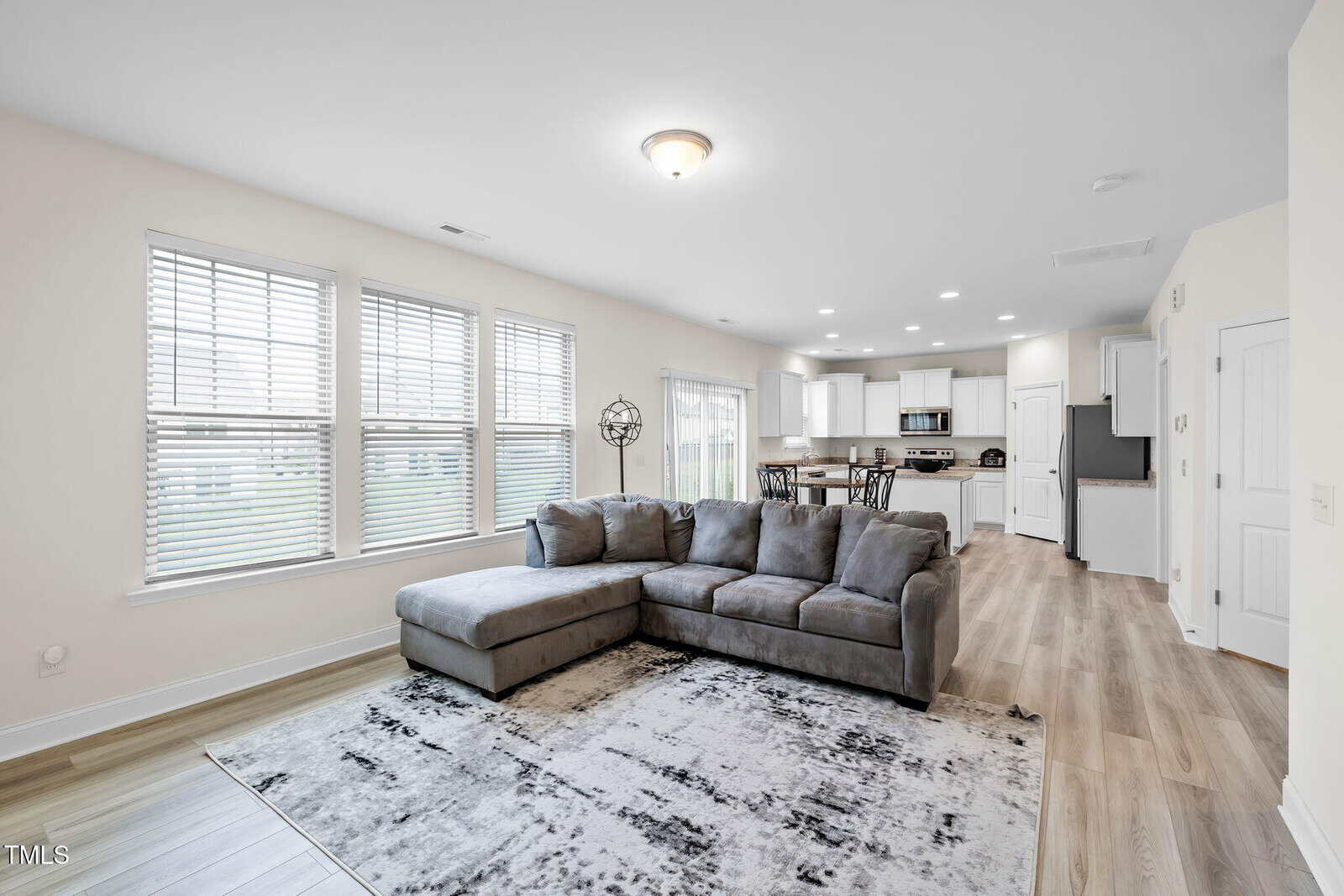 A modern living room with a gray sectional sofa and a patterned rug, adjacent to a dining area and open kitchen. Light wood flooring and large windows provide ample natural light.