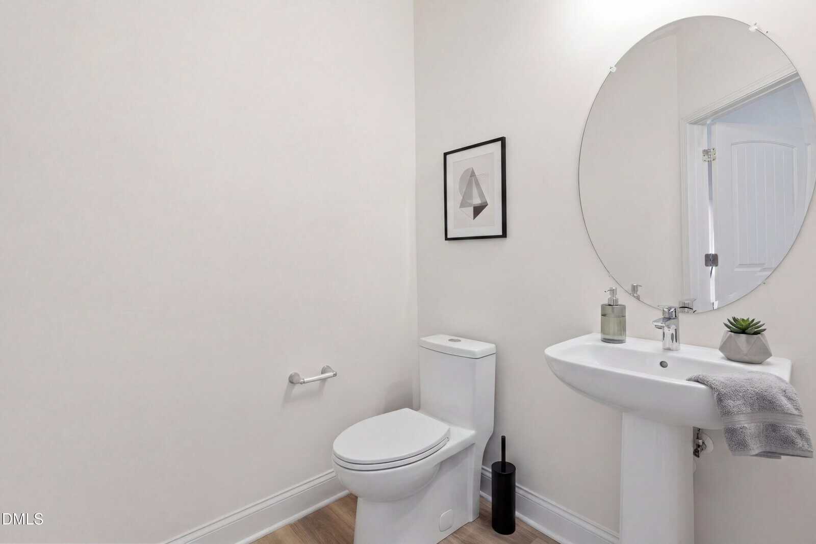A minimalist bathroom with a white toilet, pedestal sink, oval mirror, framed geometric art, a small plant, soap dispenser, and a gray hand towel on the sink. Walls and fixtures are white; floor is light wood.