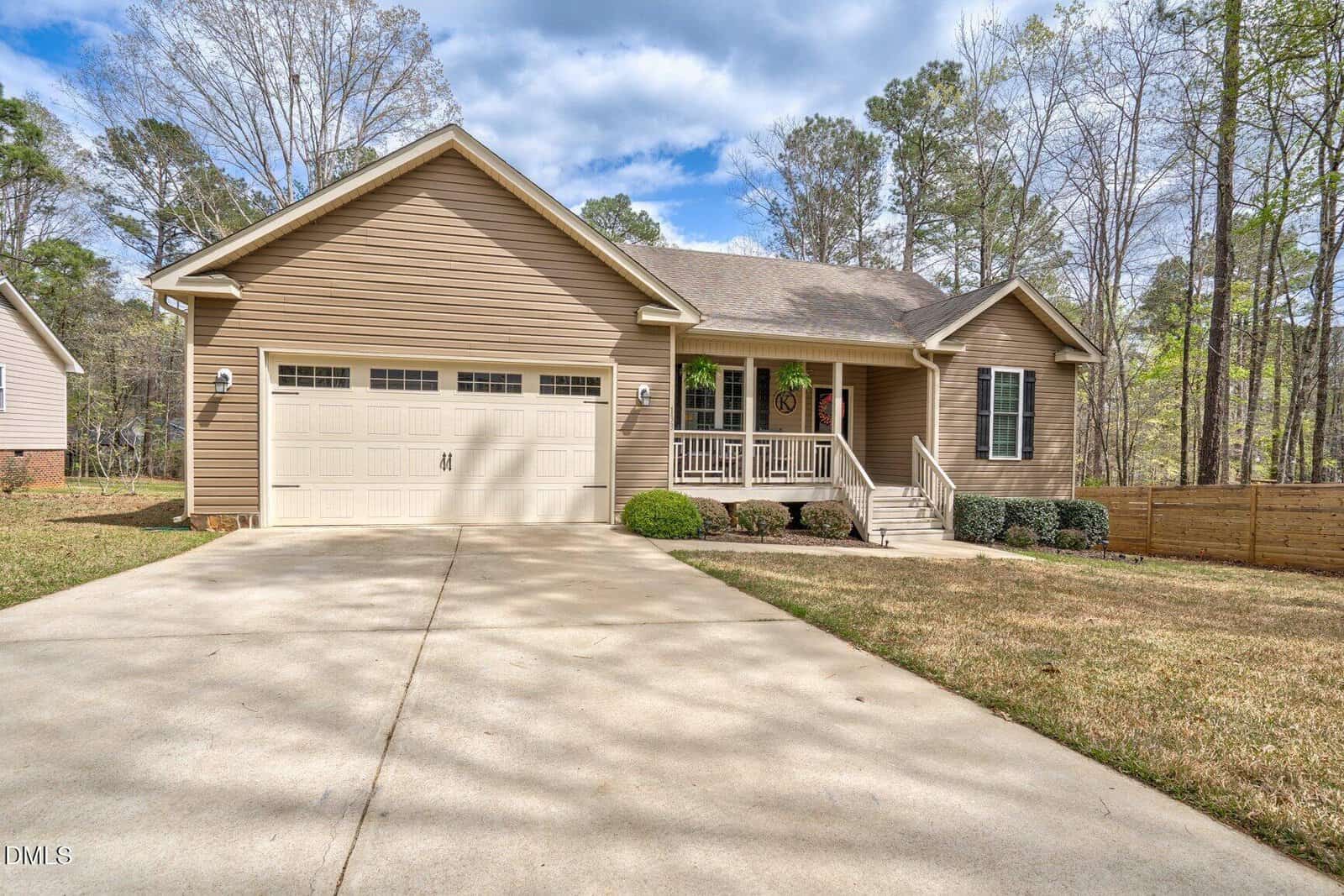 Single-story tan house with a two-car garage, covered front porch, and a wide driveway. The lawn is trimmed and trees are visible in the background. Wooden fencing runs along the right side of the property.