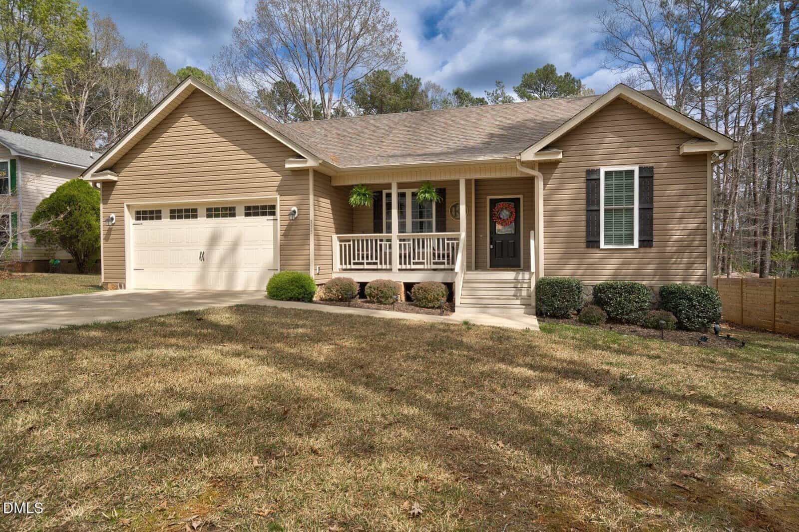 A single-story tan house with a front porch, two hanging plants, and a black front door. There is a two-car garage on the left, a concrete driveway, and a lawn with trimmed bushes in front. Trees are visible in the background.