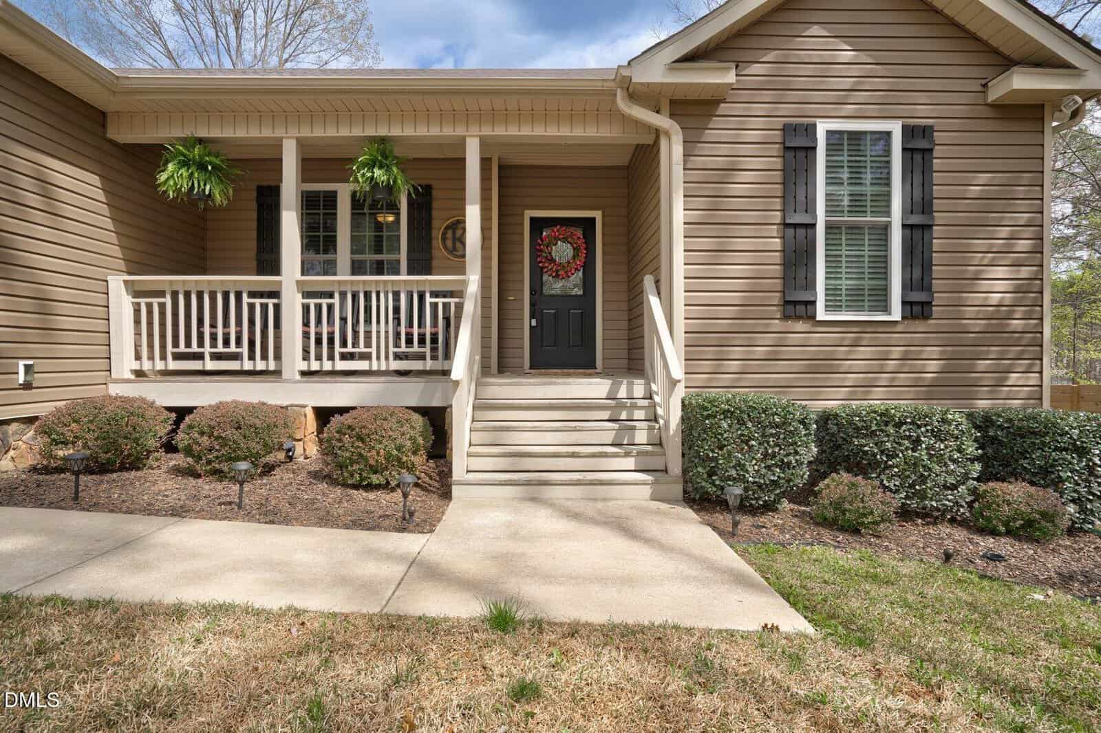 A tan, single-story house with white trim, black shutters, a covered front porch, and potted ferns. Concrete steps lead up to a black front door with a wreath. The yard has shrubs and mulch along the walkway.