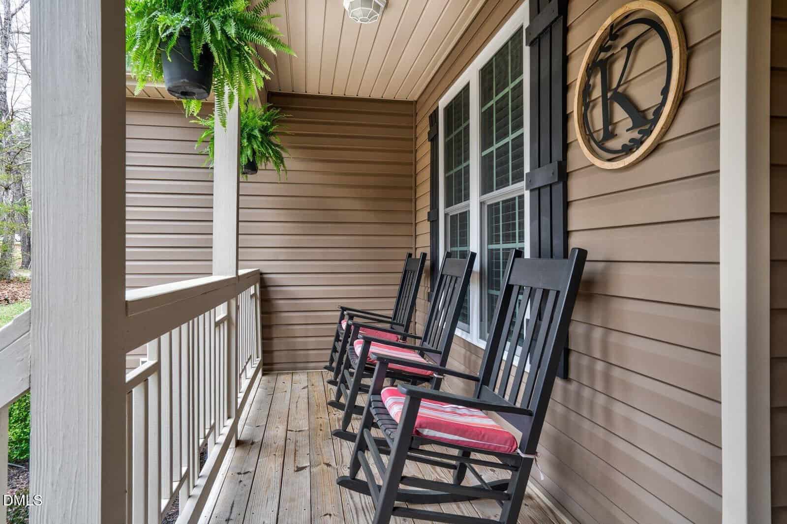 A covered porch with three black rocking chairs featuring red and white striped cushions, a hanging fern, wood railing, and light brown siding with a round wall decoration containing the letter "K.