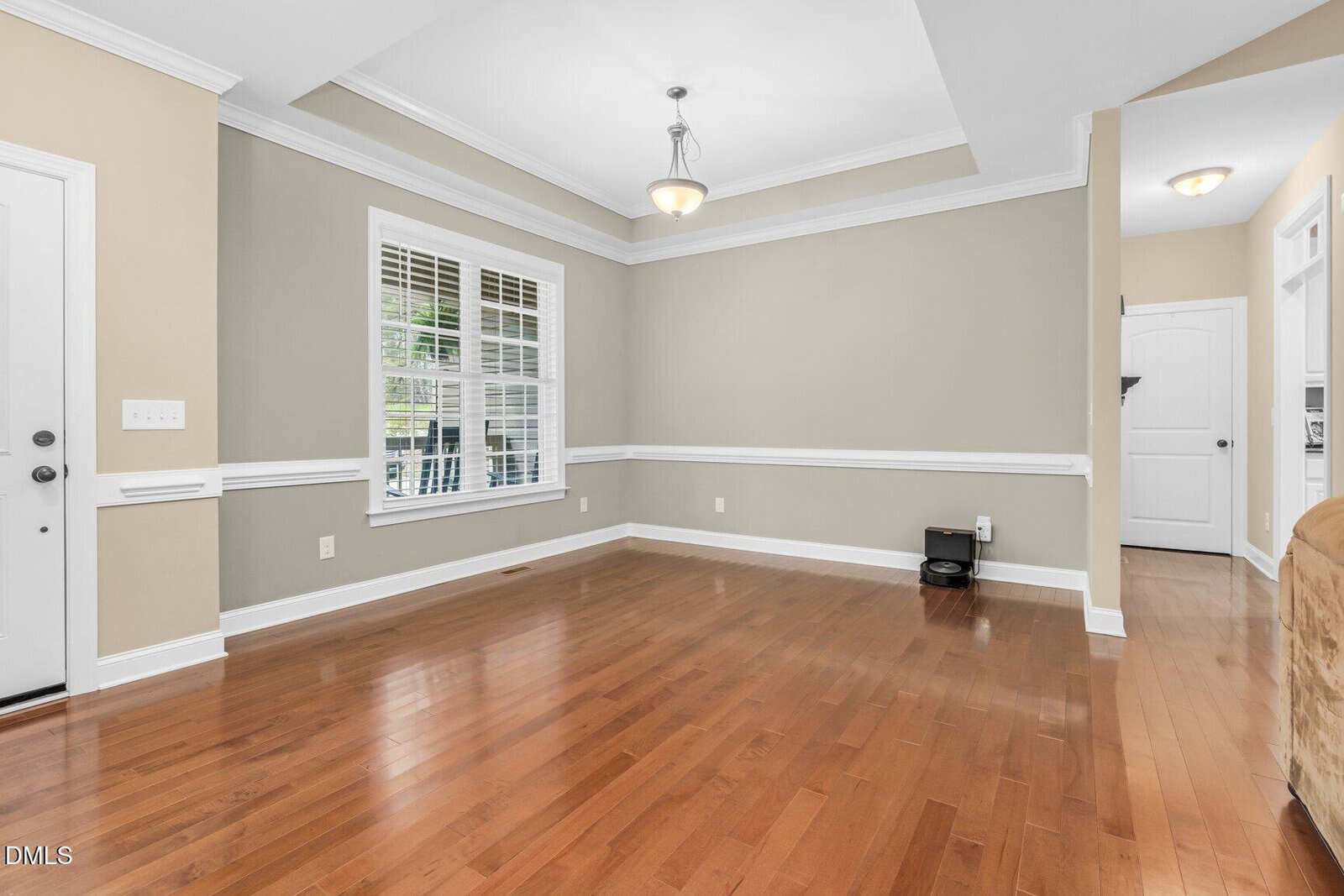 An empty room with hardwood floors, beige and light gray walls, white trim, a white framed window, a ceiling light fixture, and a white door. There is a small black robotic vacuum on the floor.