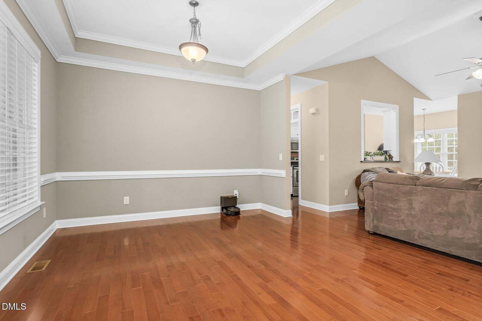A spacious, empty room with hardwood floors, beige walls, white trim, and a ceiling light. An adjoining living area with couches is visible; a robotic vacuum sits against the wall. Large windows let in natural light.