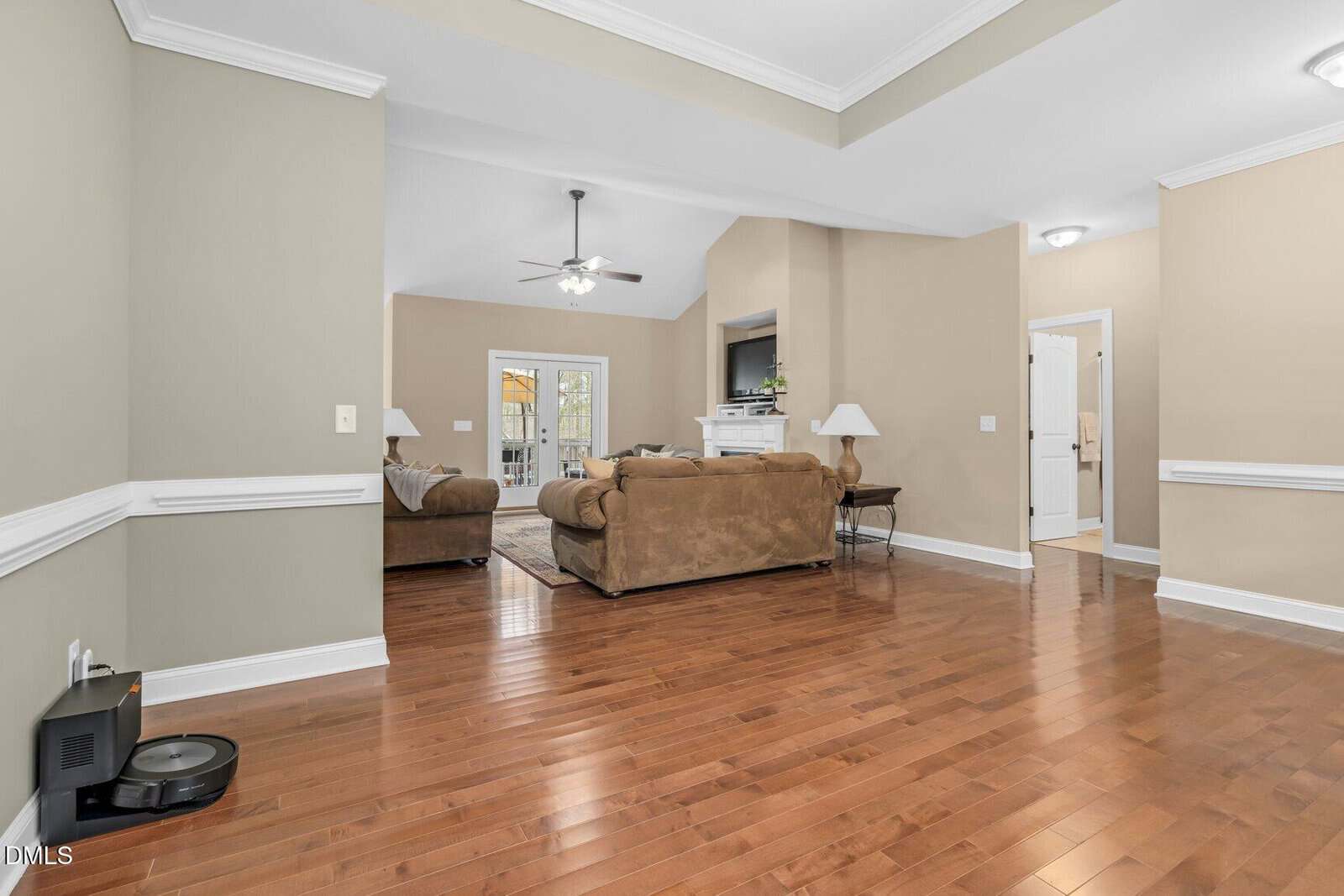 Spacious living room with tan walls, crown molding, and hardwood floors. Brown sofas face a TV and fireplace. Ceiling fan above, and double doors lead outside. A vacuum sits in the corner.