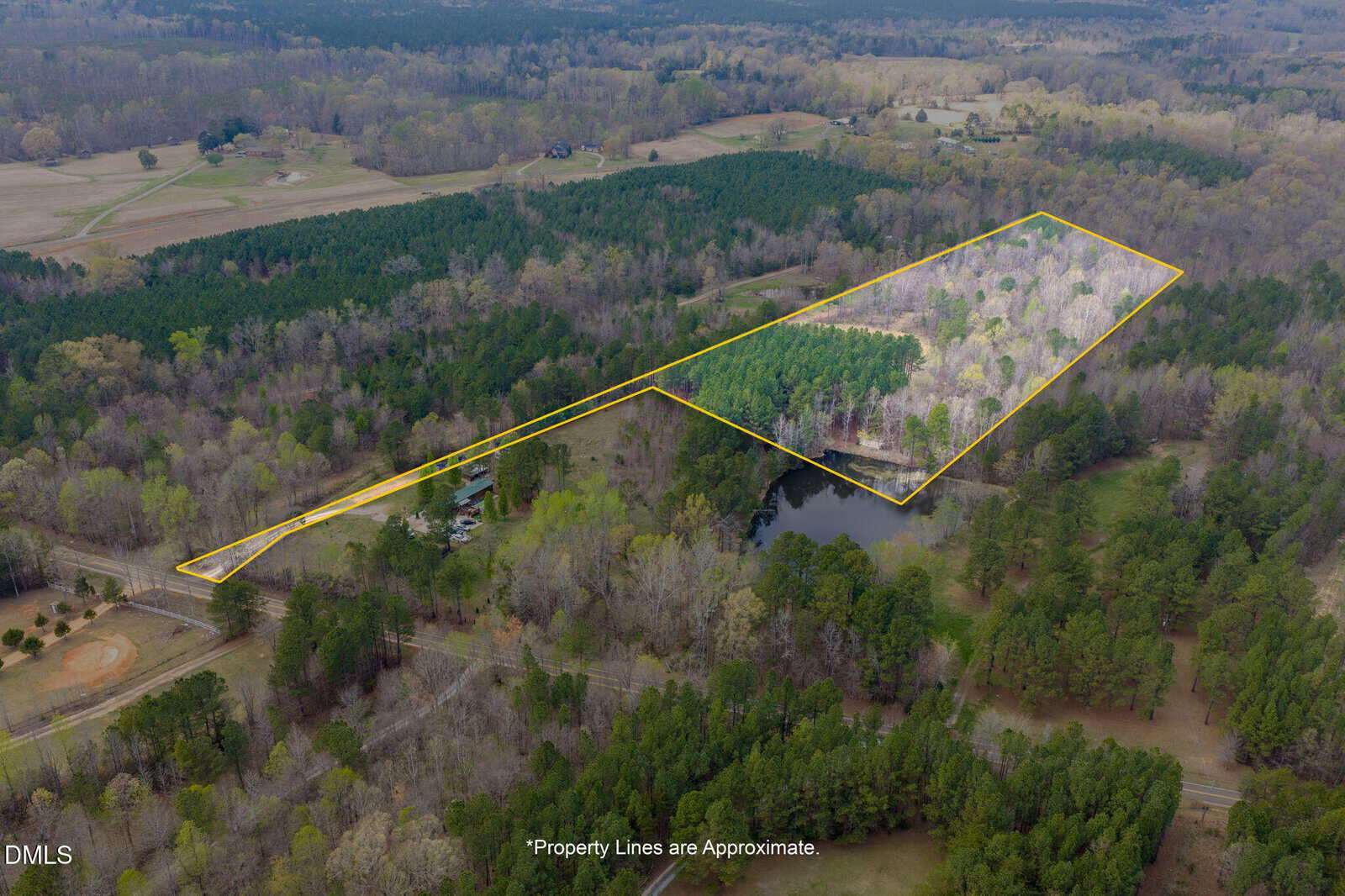 Aerial view of a wooded rural property with outlined approximate property lines, including a pond, surrounded by trees and open fields. Some houses and roads are visible in the distance.