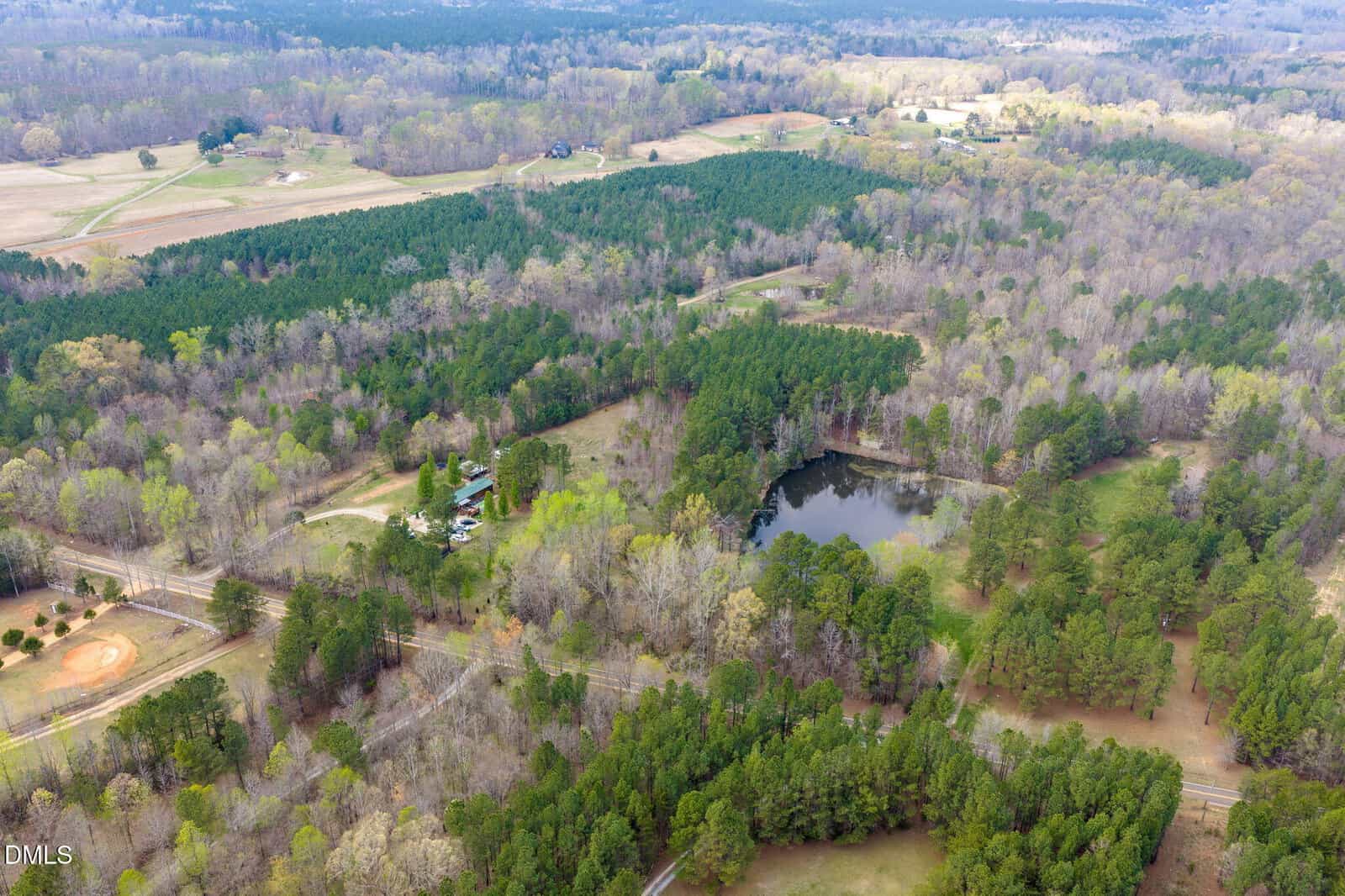 Aerial view of a rural landscape with dense trees, a pond near the center, scattered houses, open fields, and a small baseball field in the bottom left corner. Dirt and paved roads run through the area.