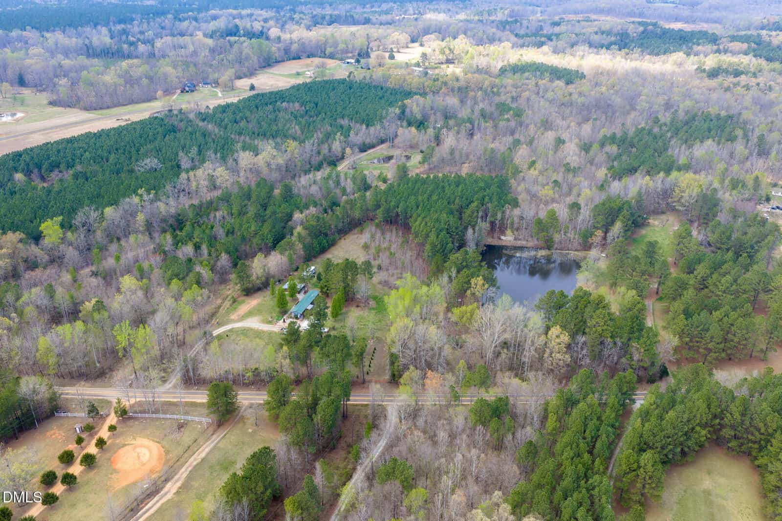Aerial view of a rural landscape with dense trees, a pond, a few houses, a baseball field, open grass areas, and intersecting roads running through the scene.
