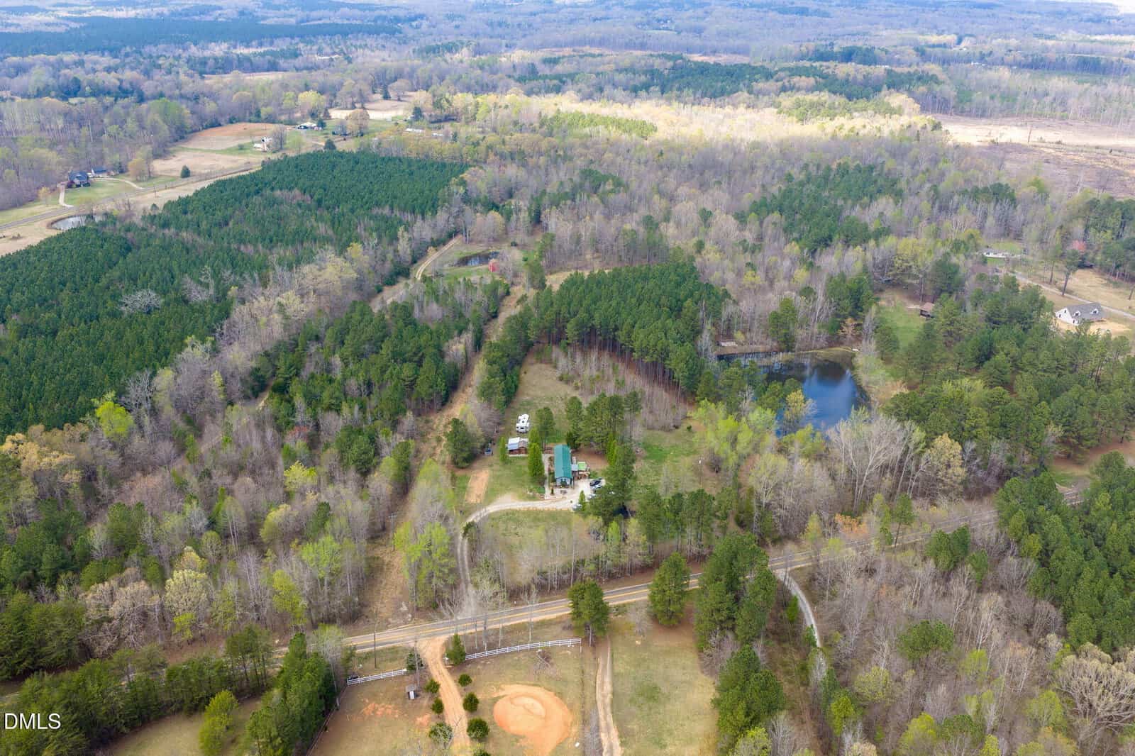 Aerial view of a rural landscape with scattered trees, a pond, a house with a few outbuildings, curved driveways, and a small baseball field; neighboring properties and forested areas are visible in the distance.