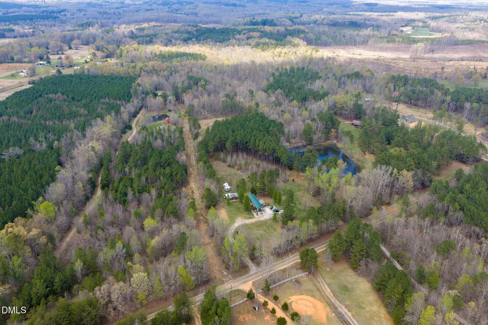 Aerial view of a rural landscape with dense trees, scattered houses, a pond, and a small sports field. Roads and open grassy areas are visible throughout the scene.