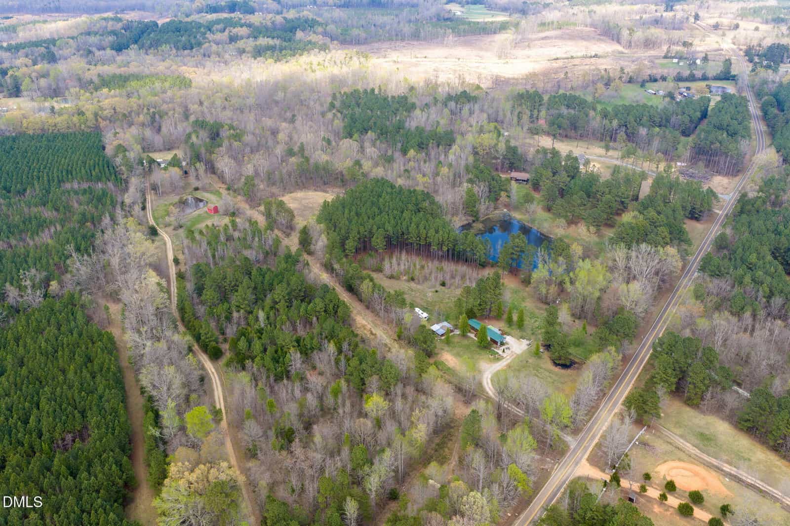 Aerial view of rural landscape with dense forest, scattered houses, a pond, winding dirt roads, a railway track, and a small baseball field. Trees are partly green, indicating early spring.
