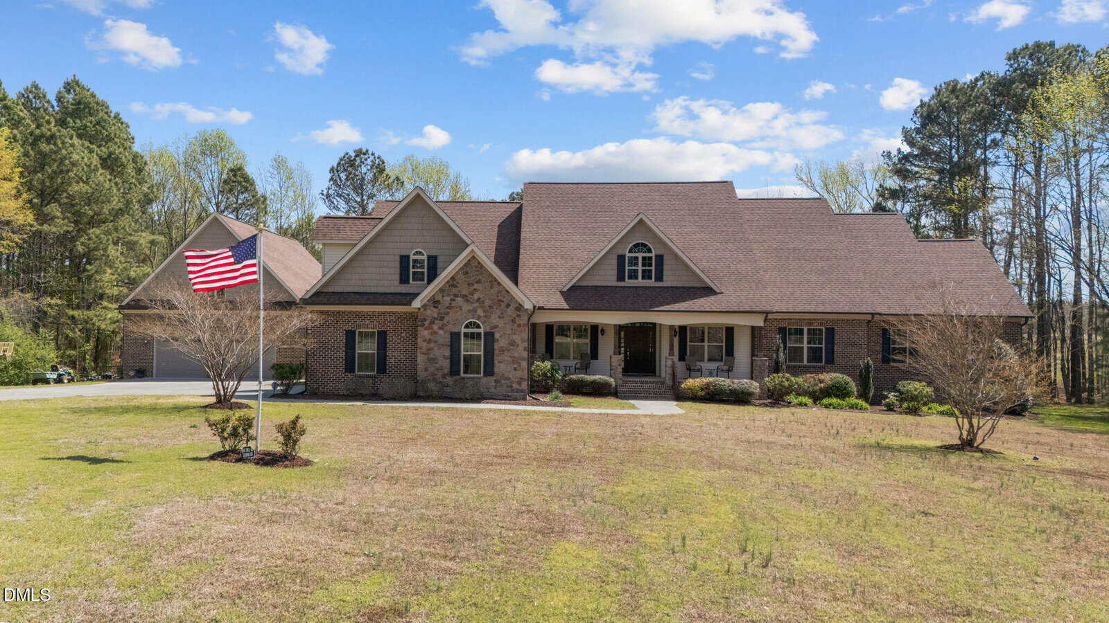 A large brick house with gabled roofs and a front porch sits on a spacious, grassy lawn. An American flag is displayed on a pole in front, and trees surround the property under a partly cloudy sky.