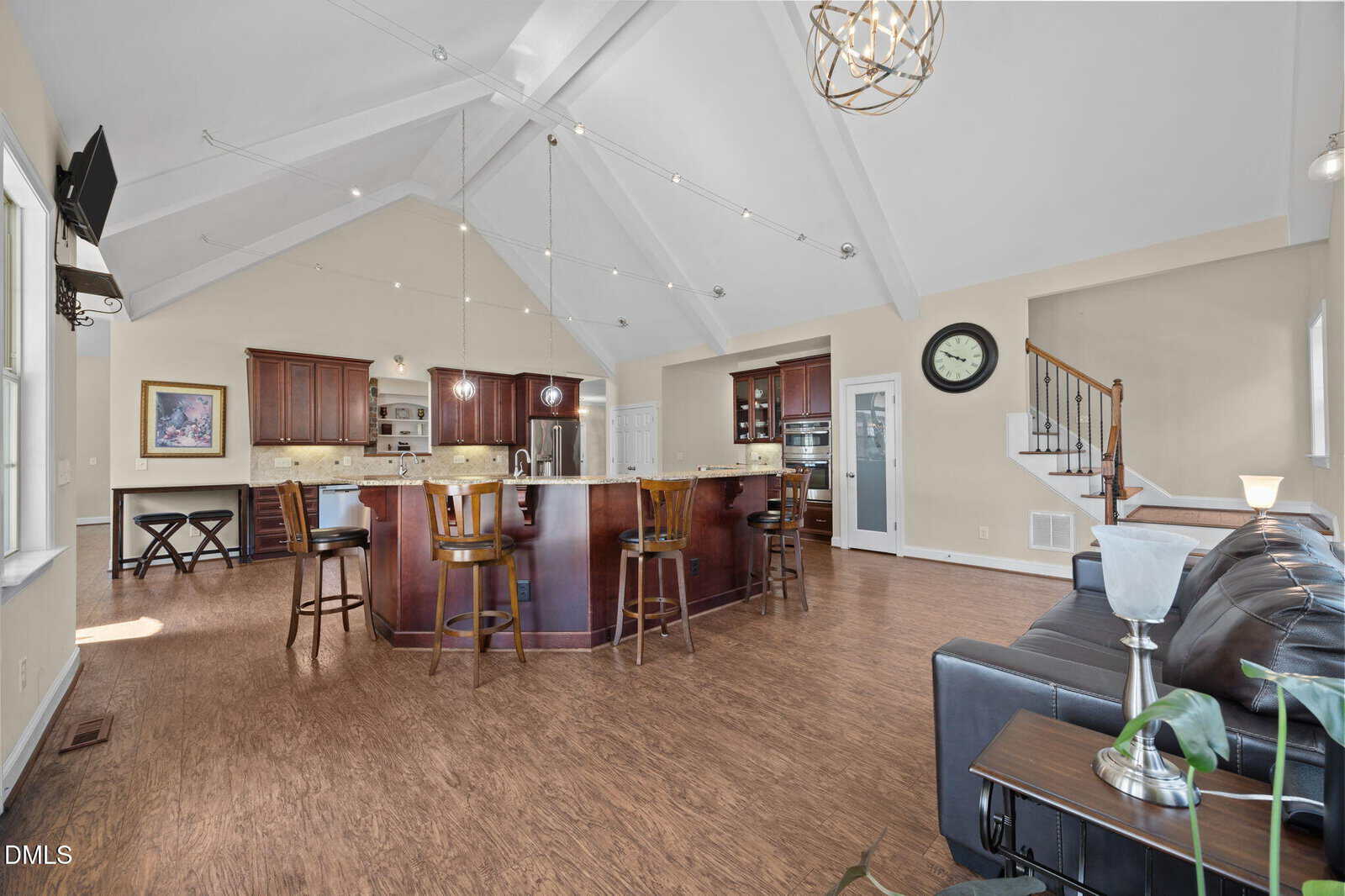 Spacious open-concept kitchen and living area with vaulted ceiling, large island with barstools, dark wood cabinets, a black leather sofa, wall clock, and natural light from several windows.