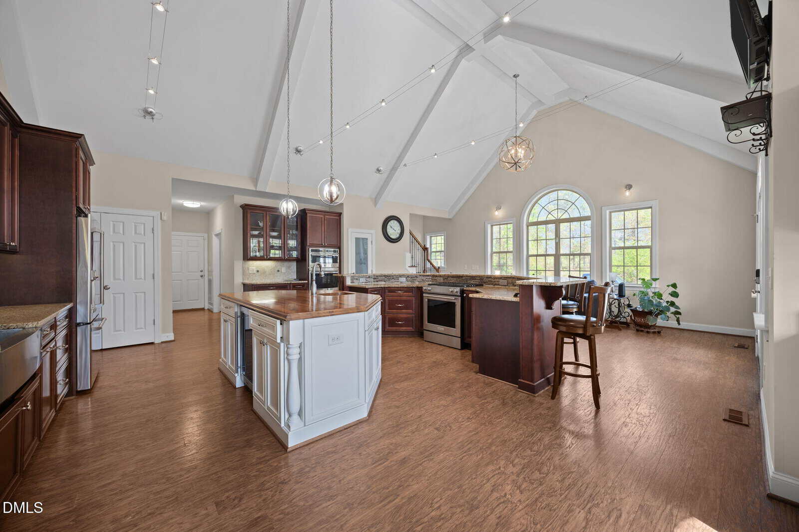 Spacious kitchen with vaulted ceiling, large arched windows, wood floors, dark wood cabinets, a white island with pendant lights, and bar stools. Natural light fills the room, and a plant sits by the windows.