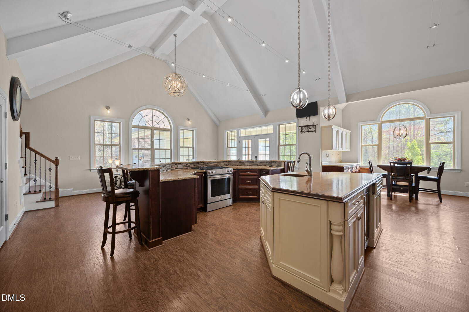 Spacious kitchen with vaulted ceiling, island, stainless steel appliances, wood flooring, and large arched windows allowing natural light. Dining area in the corner near windows; staircase visible on the left.