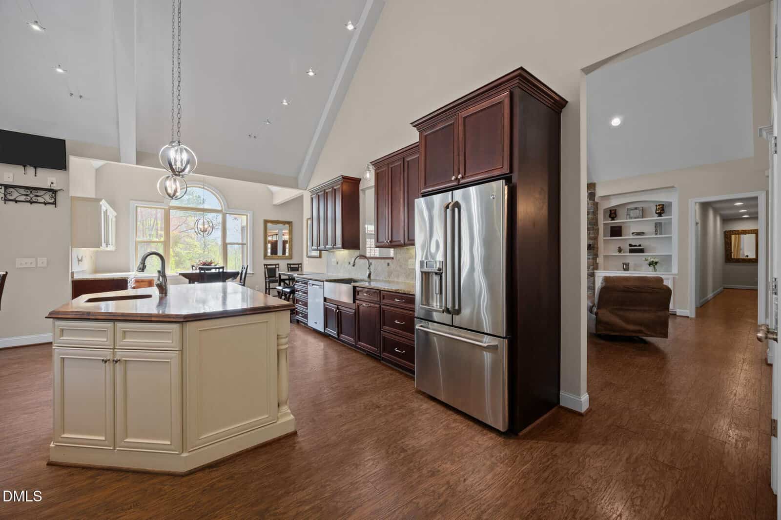 Spacious kitchen with dark wood cabinets, stainless steel appliances, large island with sink, and open view into a living room area with built-in shelves. Natural light streams in through a large arched window.