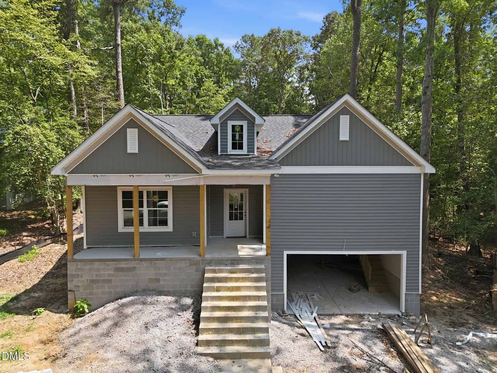 A gray house under construction with a covered front porch, wooden posts, concrete steps, and an unfinished garage, surrounded by trees and dirt.