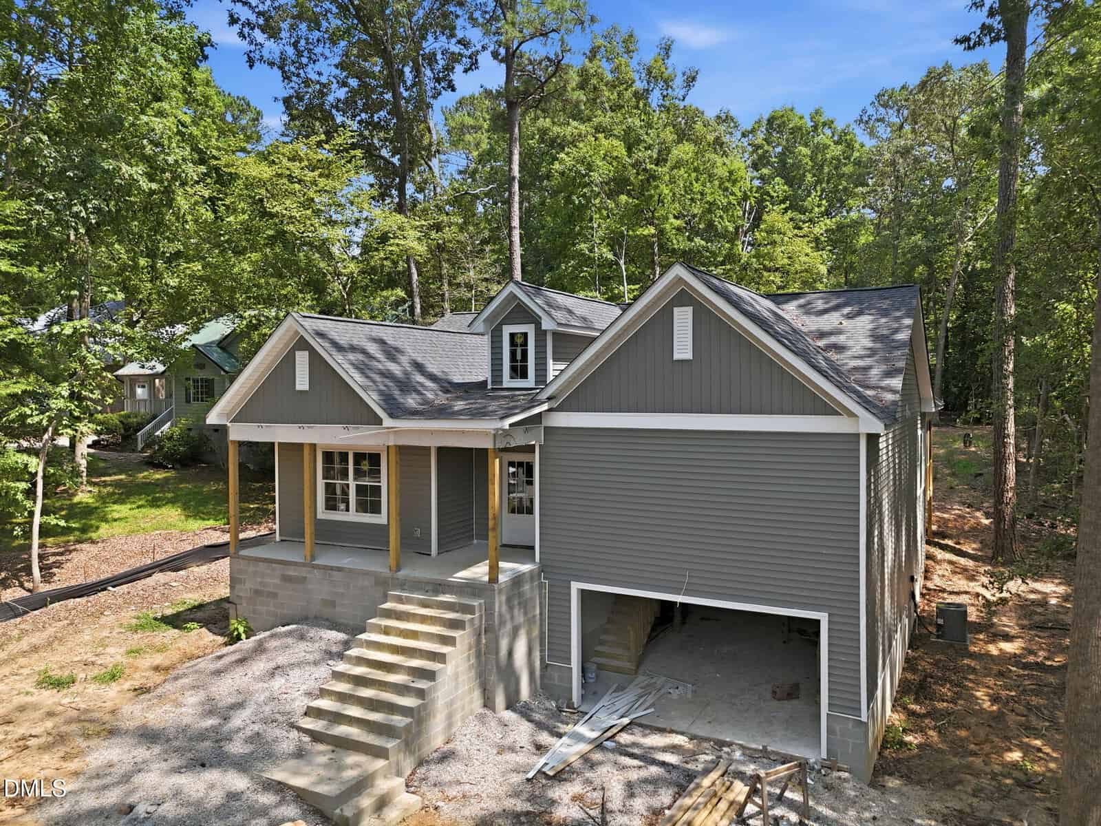 Gray house with a front porch, white trim, and attached two-car garage under construction, surrounded by trees on a sunny day. Steps lead from the yard to the porch, and building materials are visible in the driveway.