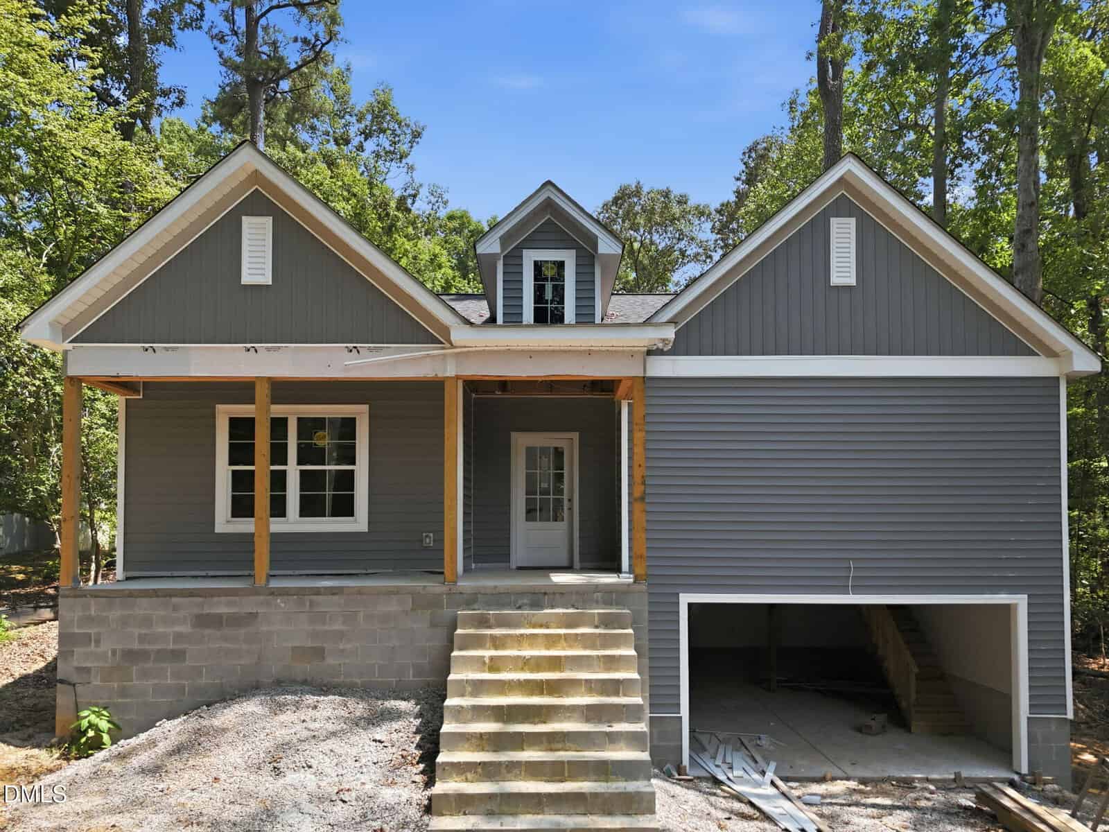 Gray house under construction at 102 Mayan Drive, Louisburg, with a front porch, steps leading to the main door, and an unfinished garage underneath. Surrounded by trees, the house features white trim and an upper dormer window.