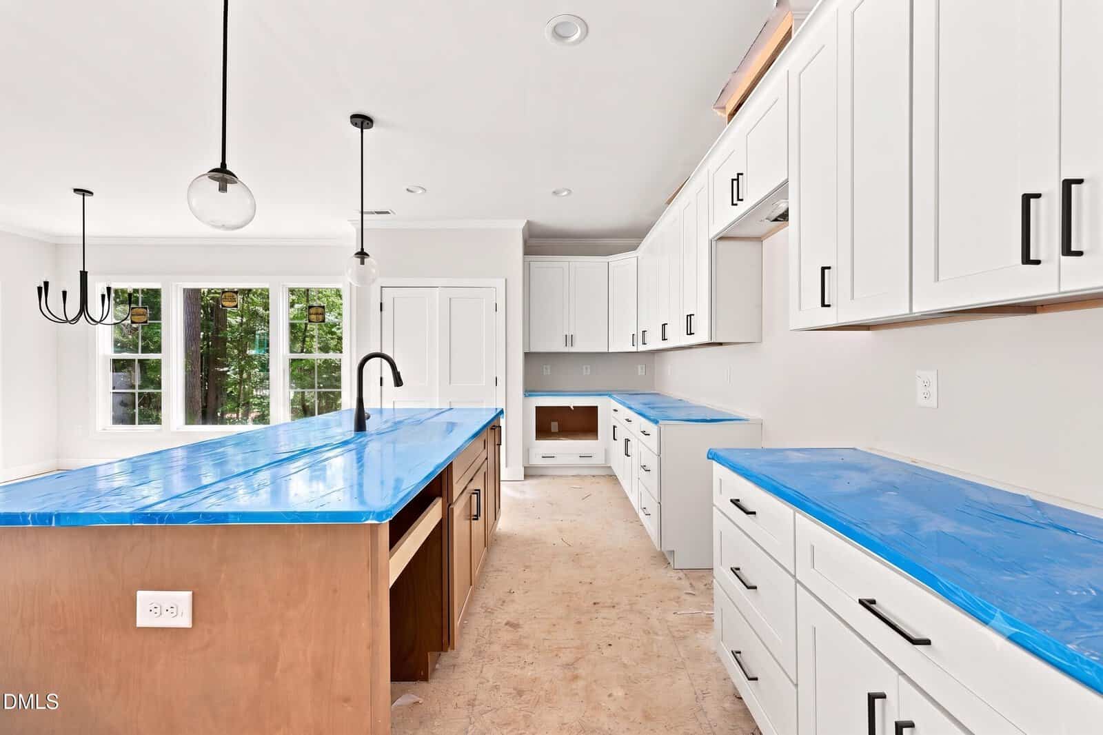 Bright kitchen with white cabinets, black handles, two islands covered in blue protective wrap, pendant lights, large windows, and unfinished floor.
