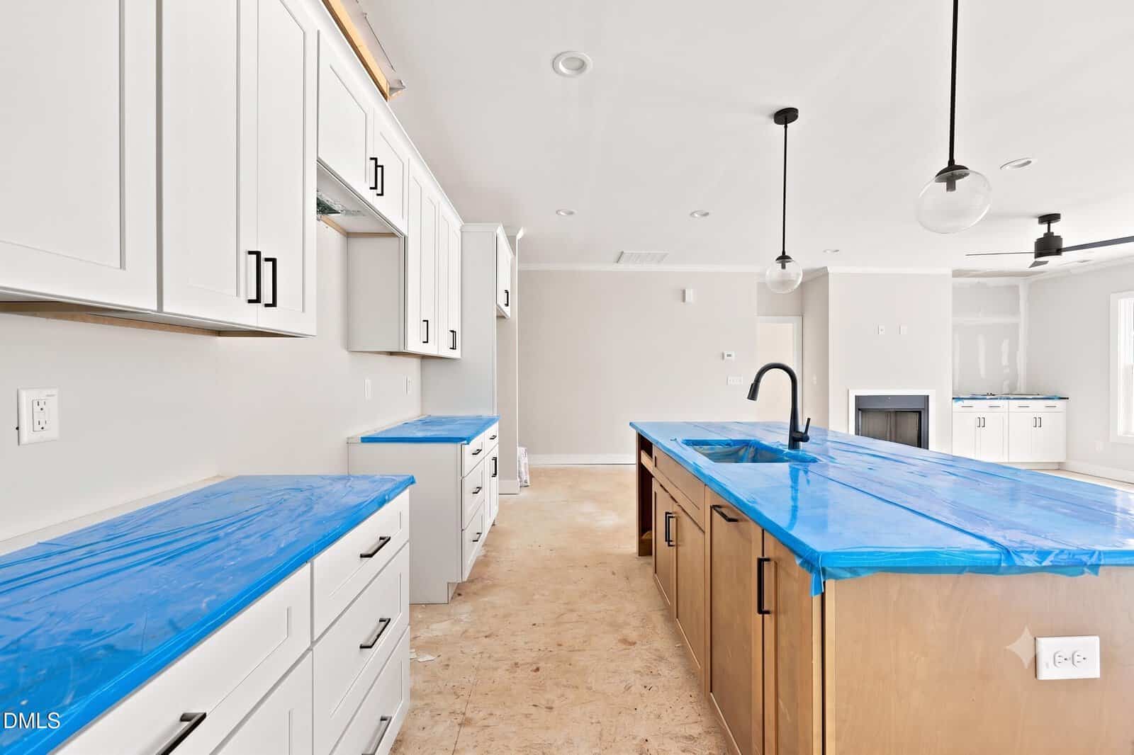 A modern kitchen under construction with white cabinets, a wood island, and blue protective plastic covering the countertops. Light fixtures and unfinished flooring are visible.