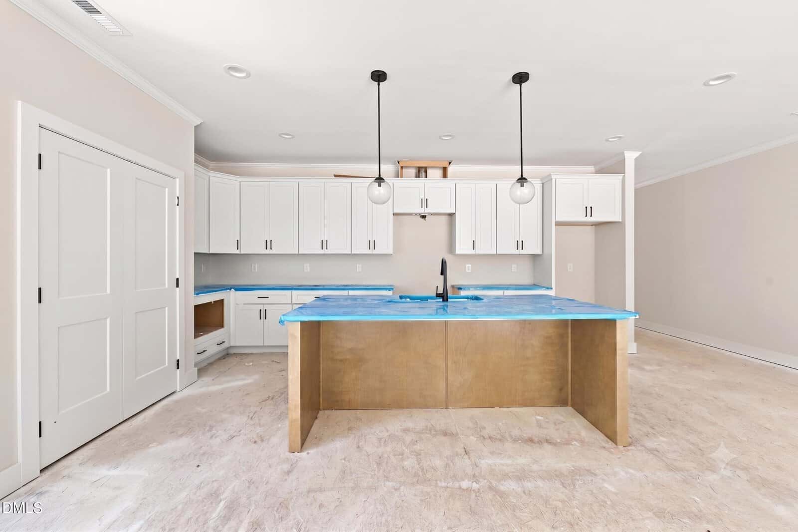 Modern kitchen under construction with white cabinets, a central island with a gold base, blue protective coverings on countertops, and two pendant lights hanging above the island. The floor is unfinished.