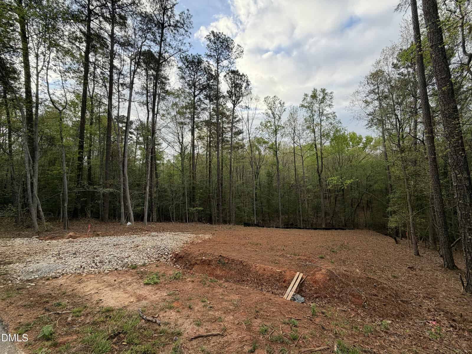 A cleared patch of reddish dirt and gravel sits in a wooded area with tall trees. The ground is uneven with a small trench and wooden planks, suggesting site preparation or early construction work.