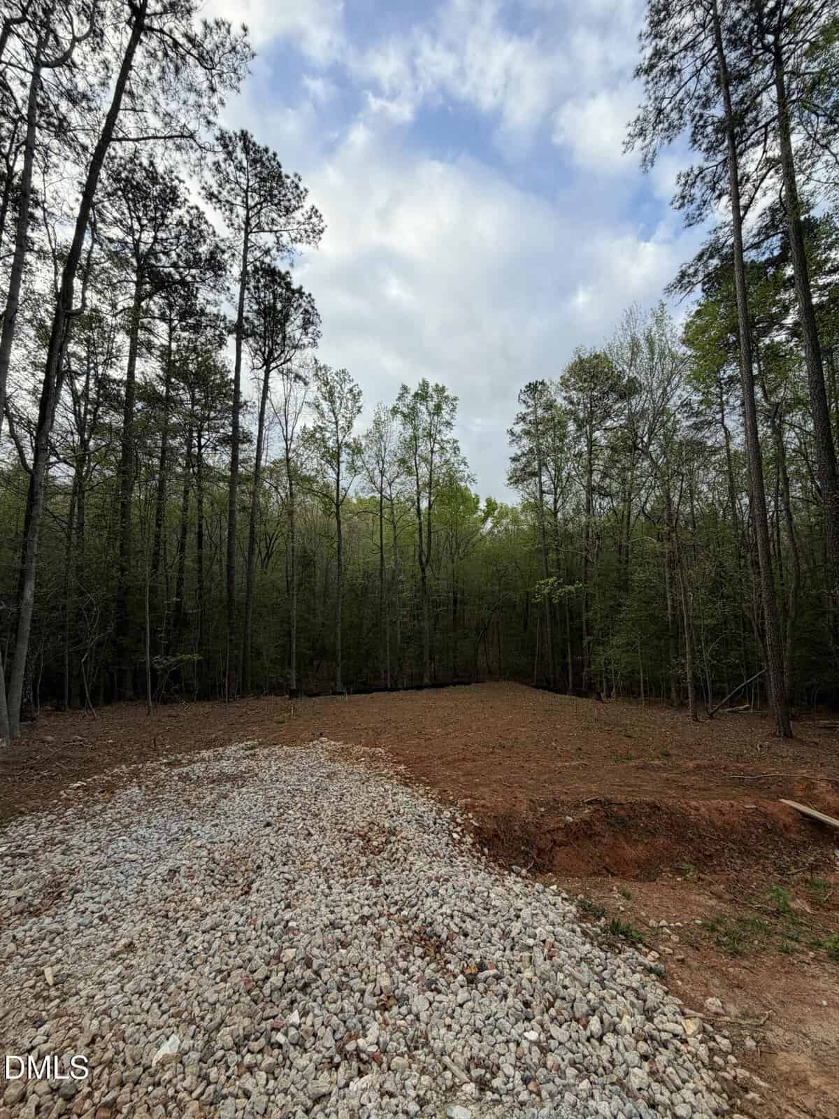 A cleared plot of land bordered by tall trees, with a section of gravel in the foreground and an area of bare soil. The sky above is partly cloudy with patches of blue.