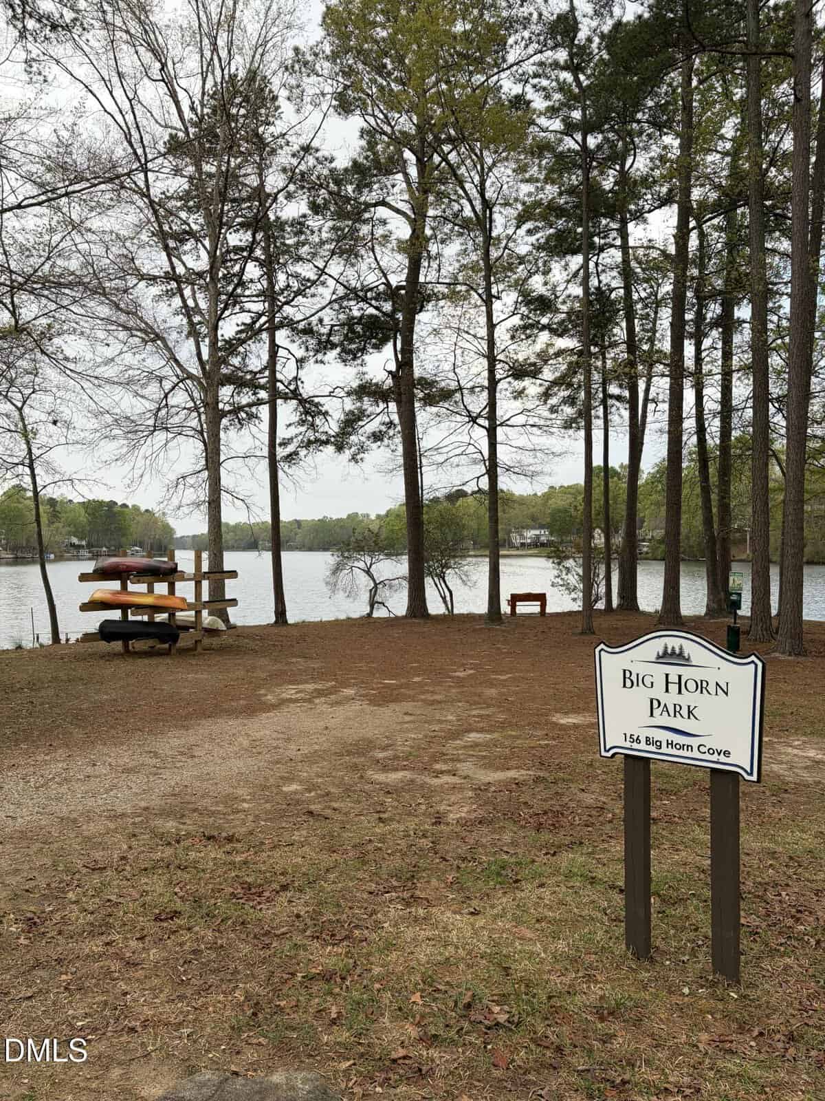 A lakeside park with tall trees, picnic tables, and a sign that reads “Big Horn Park, 154 Big Horn Cove.” Calm water and houses are visible in the background.