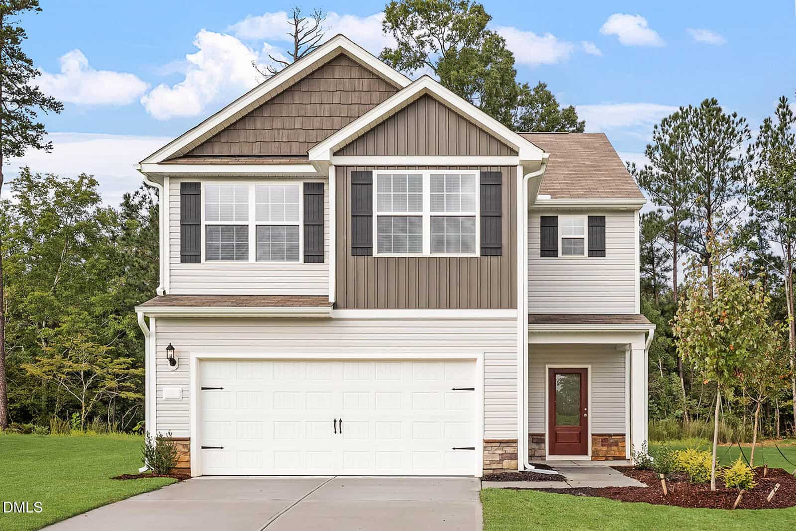 A two-story suburban house with gray and white siding, black shutters, a double garage, a red front door, and a driveway. The yard has green grass, a small tree, and is surrounded by trees in the background.