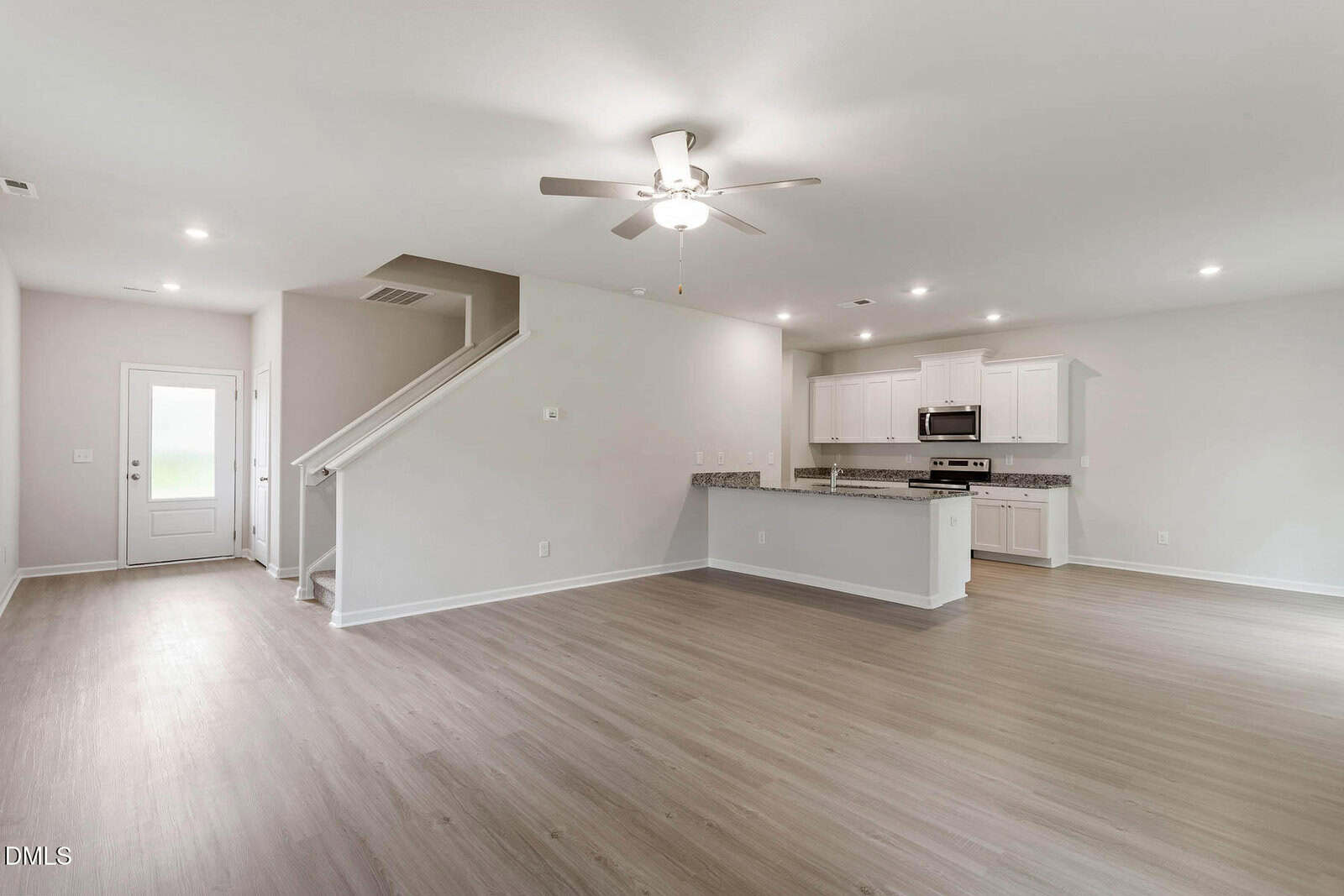 Open-plan living area with light wood flooring, white walls, and recessed lighting. A staircase leads upstairs, and a modern kitchen with white cabinets and granite countertops is visible in the background.