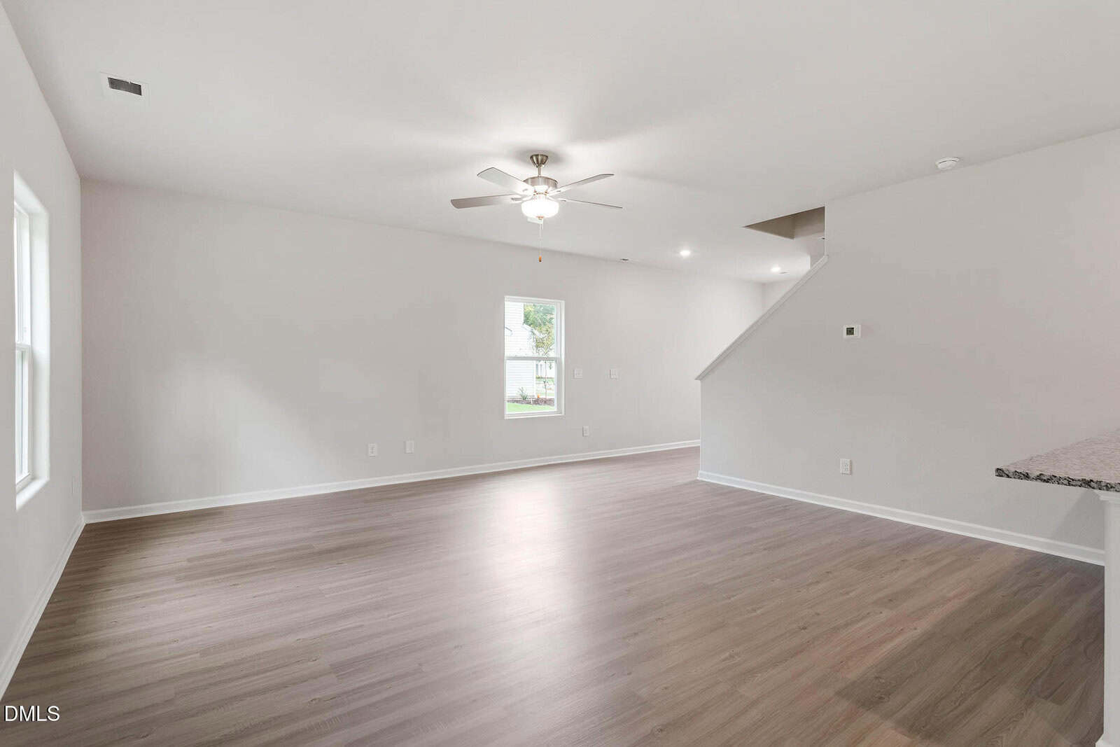 Empty living room with light wood flooring, white walls, a ceiling fan with lights, a window, and partial view of a staircase and kitchen countertop on the right. Natural light enters through the window.
