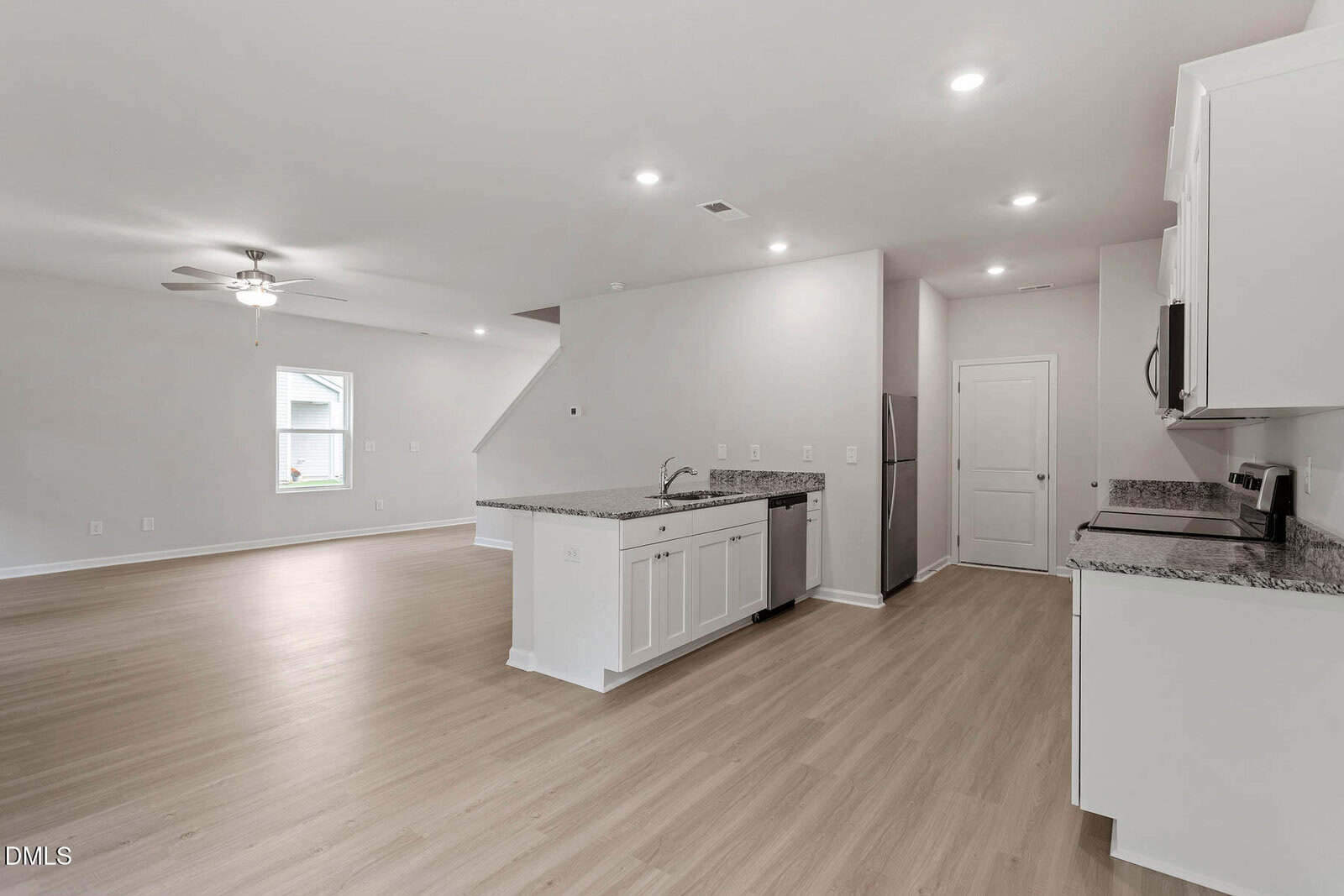 Open-concept kitchen and living area with light wood floors, white cabinets, granite countertops, recessed lighting, ceiling fan, and a window providing natural light. The space has a modern, minimalistic design.