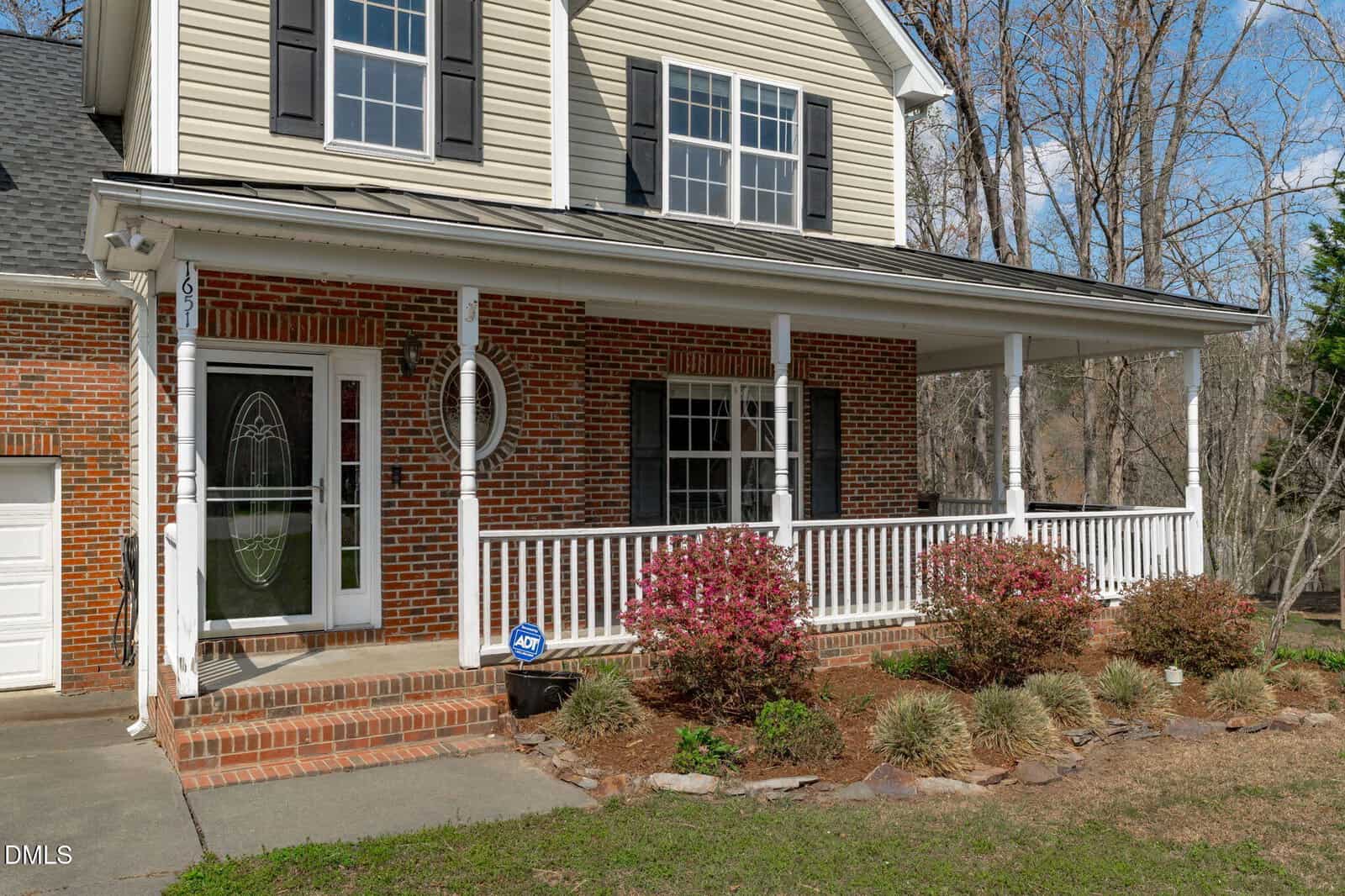 A brick house with white trim, black shutters, a covered front porch, railing, bushes, and flowers in the yard. The porch features columns and an oval glass front door. Trees without leaves are visible in the background.