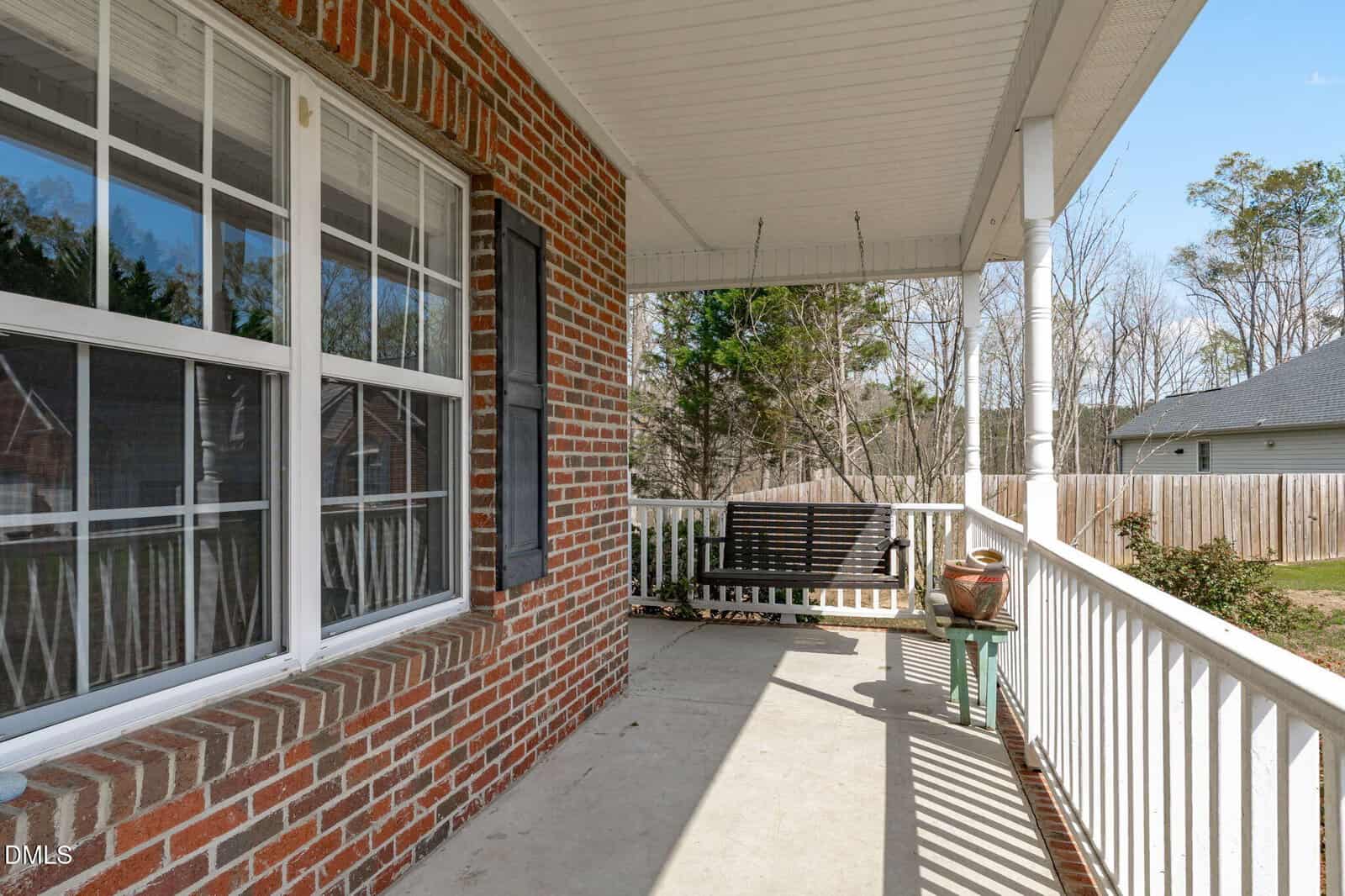 A brick house porch with white railing, a hanging wooden bench, potted plants, and trees visible in the background on a sunny day.