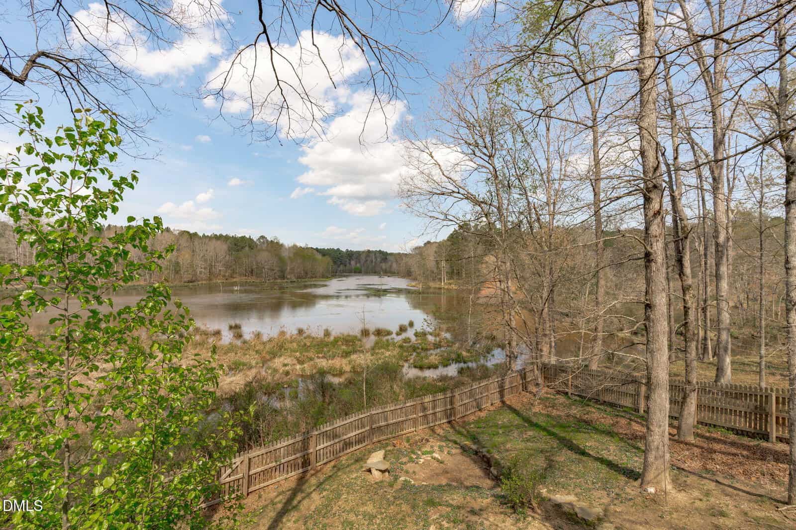 View of a calm lake surrounded by trees, some with early spring leaves. A wooden fence runs along the yard in the foreground, and the sky is bright with scattered clouds.
