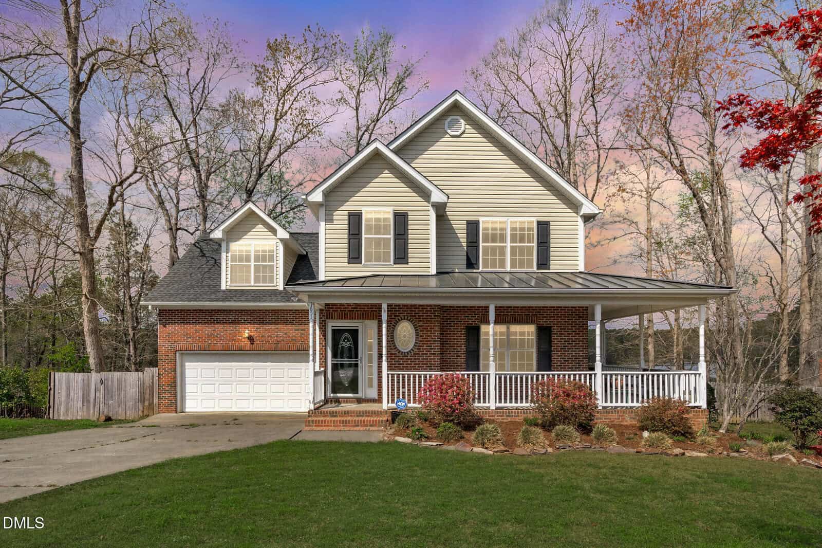 Two-story suburban house with beige siding and red brick exterior, white trim, double garage, covered front porch, and a manicured lawn, set against a backdrop of tall trees and a colorful sunset sky.
