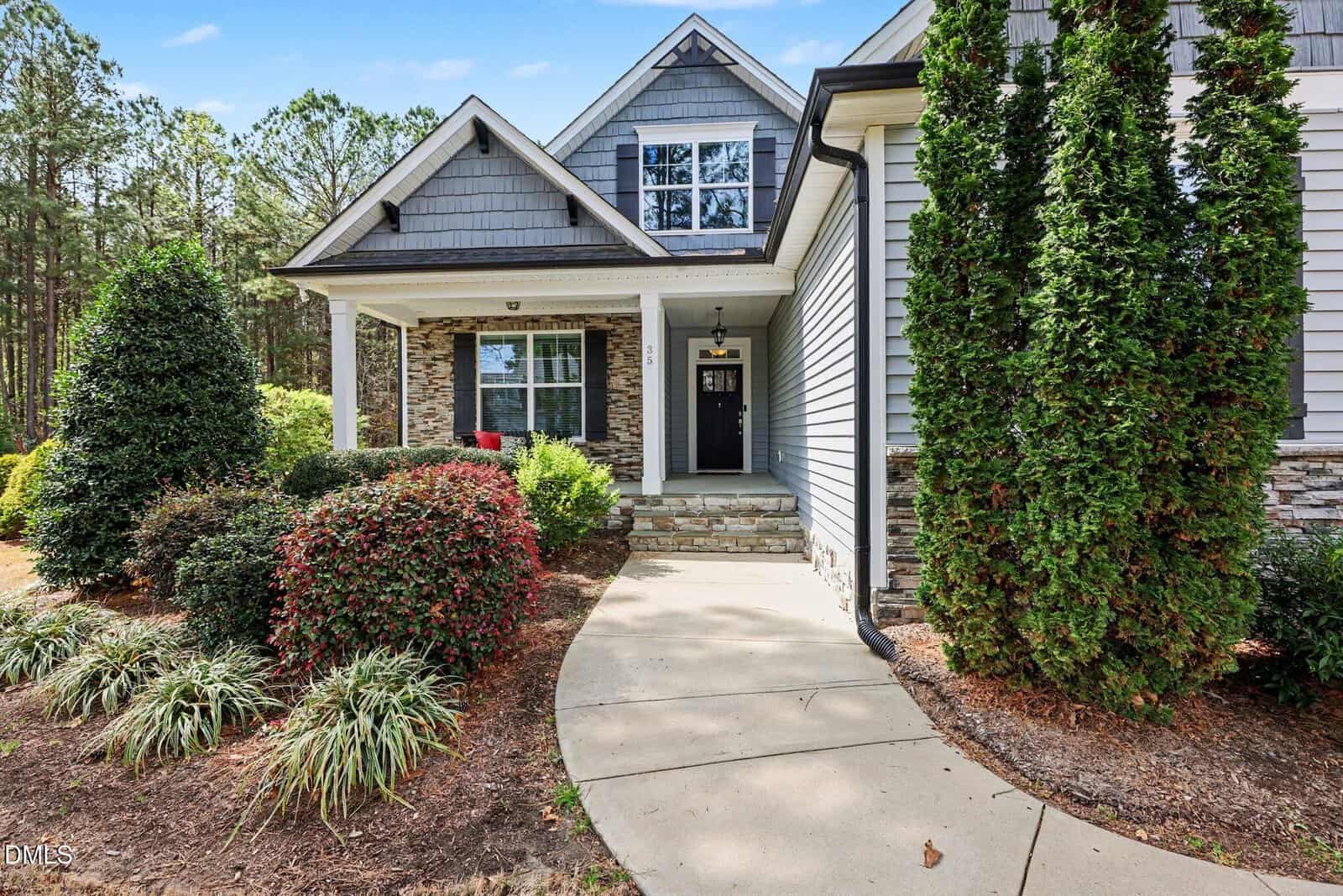 A concrete walkway curves toward the covered front porch of a gray house with stone accents. Bushes and trees line the path, and a lawn with mulch is visible in the landscaped front yard.