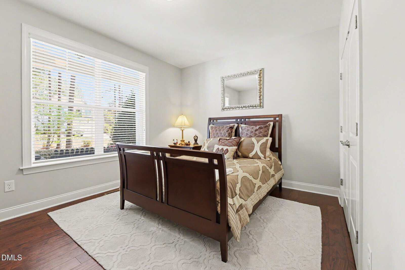 Bright bedroom with a wooden bed, patterned bedding, white rug, nightstand with lamp, and a decorative mirror above the bed. Large window lets in natural light. Walls are light-colored.