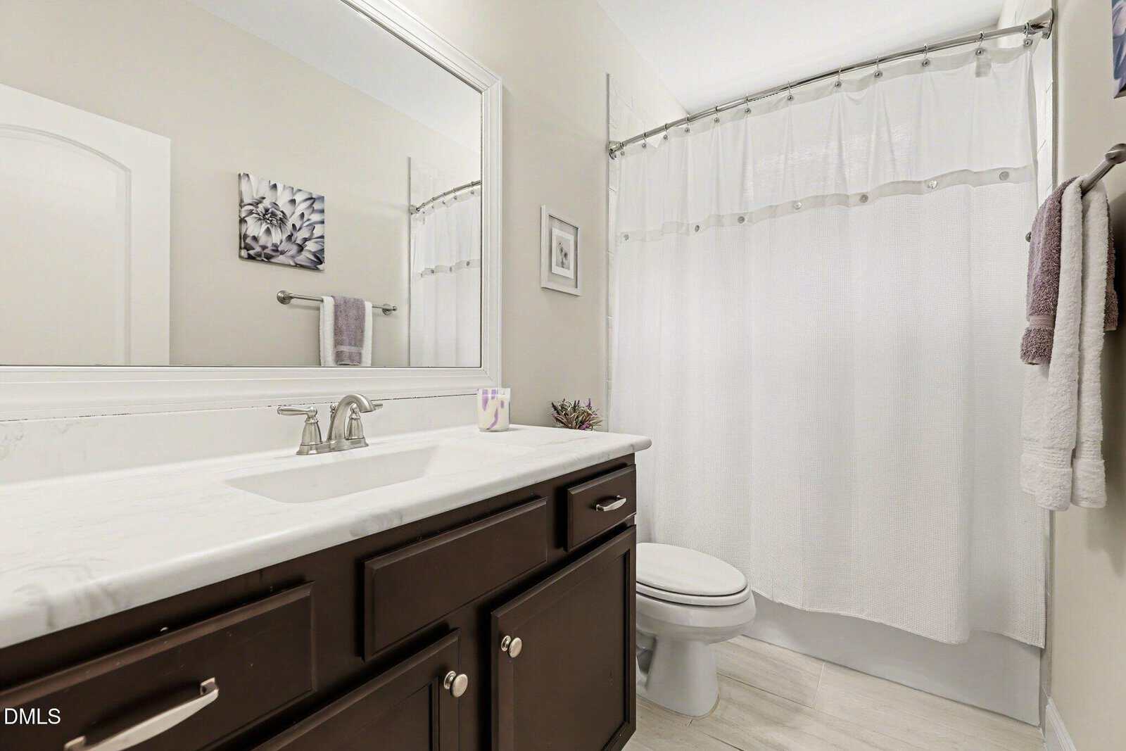 A modern bathroom with dark wood cabinets, a white countertop, large mirror, toilet, and a white shower curtain. Towels hang on a rack, and the wall features floral artwork.