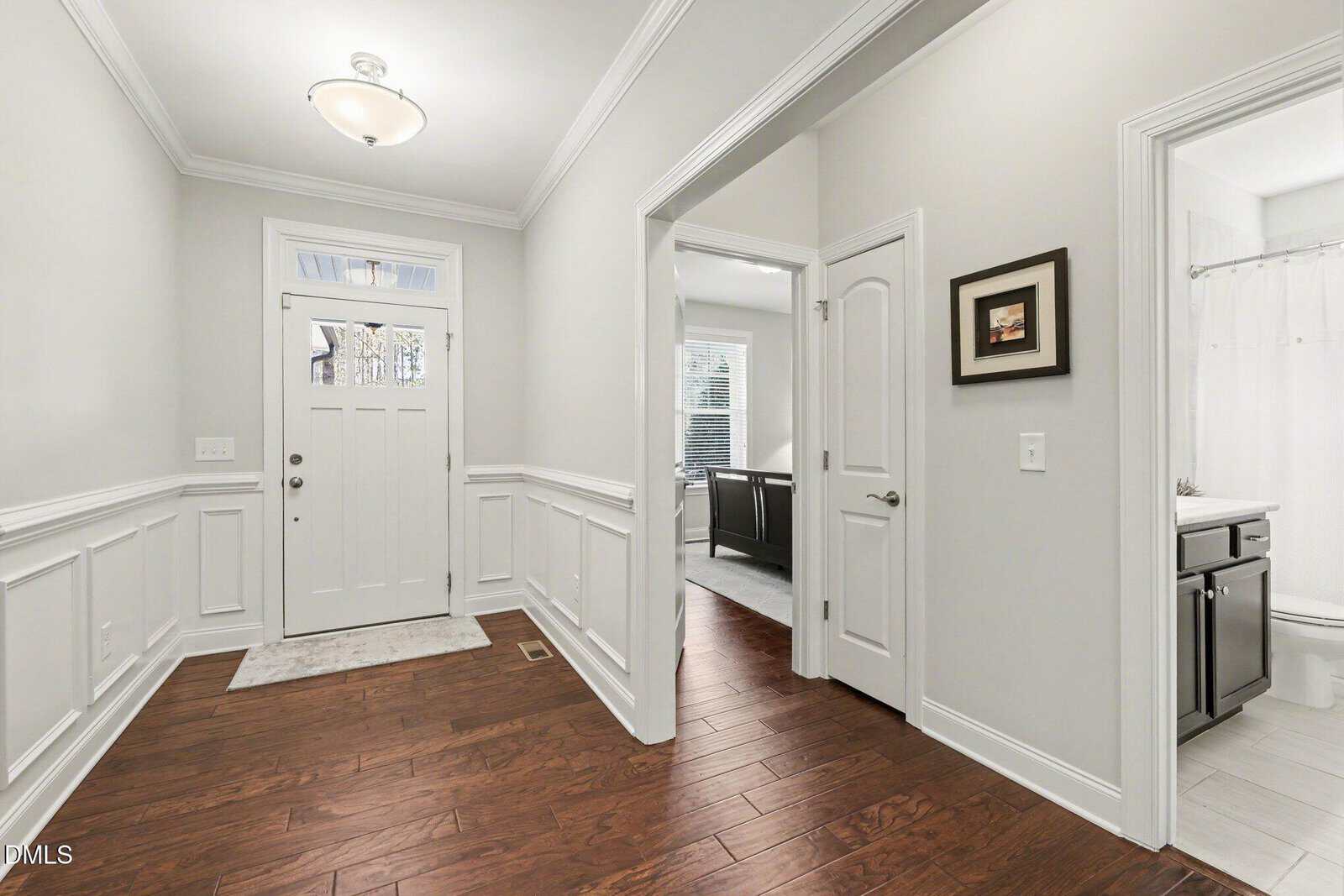 A hallway with wooden floors, white wainscoting, and light gray walls. There is a white front door with glass panels, two open doorways, a closed door, ceiling light, and framed art on the wall.