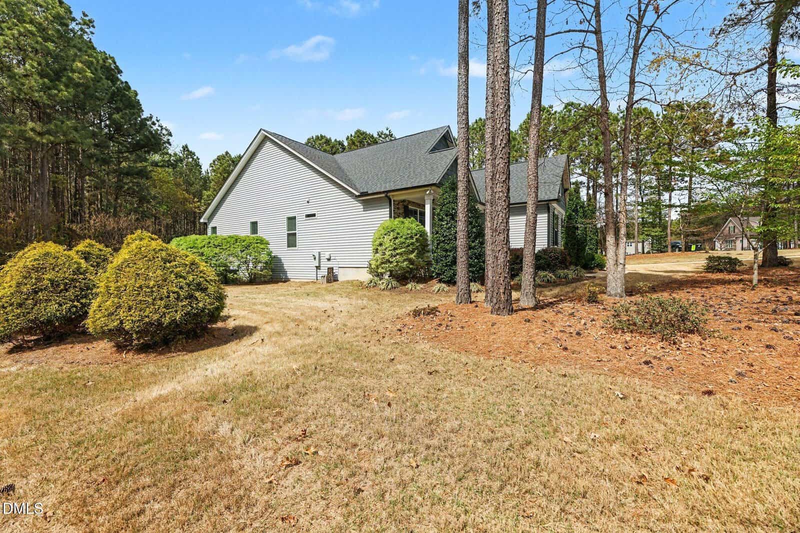 A single-story house with light siding and a dark roof is surrounded by trees and shrubs. The yard has dry grass and pine straw, with a forested area in the background under a clear sky.