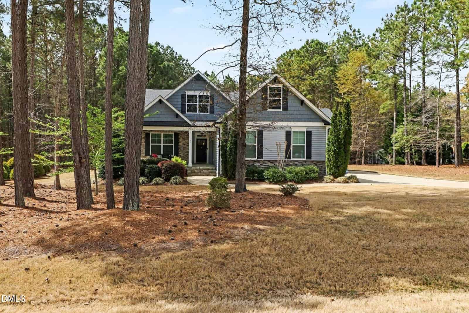A two-story house with grey siding and white trim is surrounded by trees and natural landscaping, with a driveway on the right and a dry, brown lawn in front.