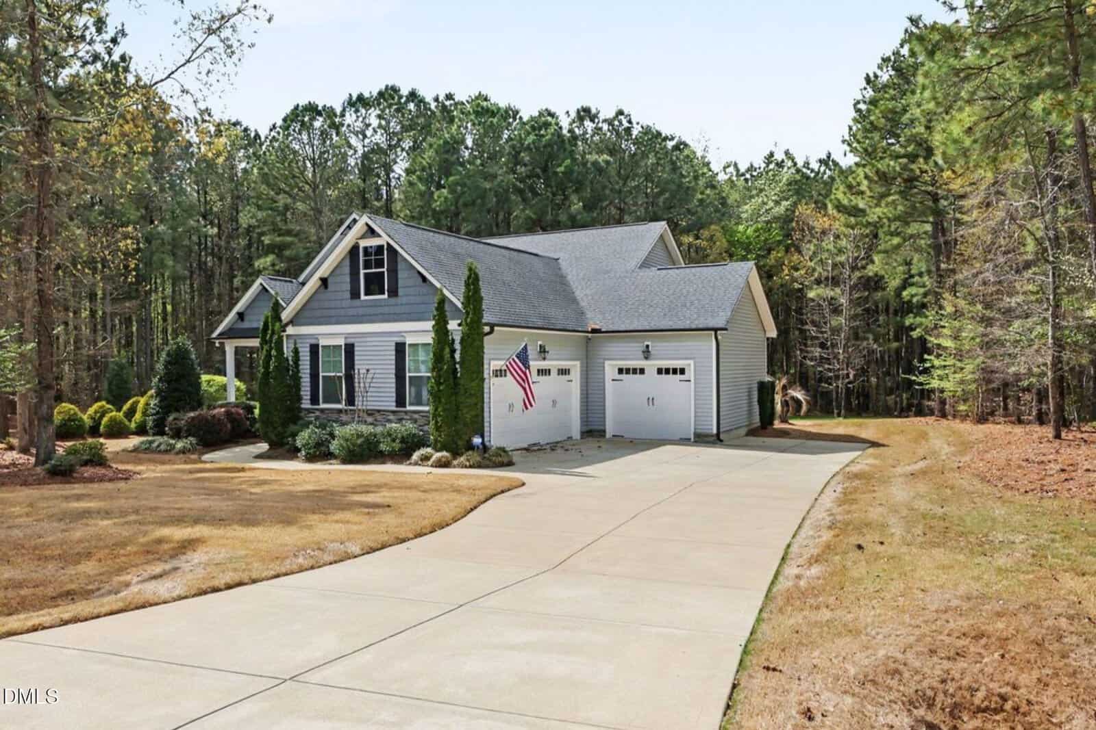 A single-story house with light-colored siding, an American flag, two garage doors, and a wide driveway, surrounded by trees and a lawn with some shrubs and plants.