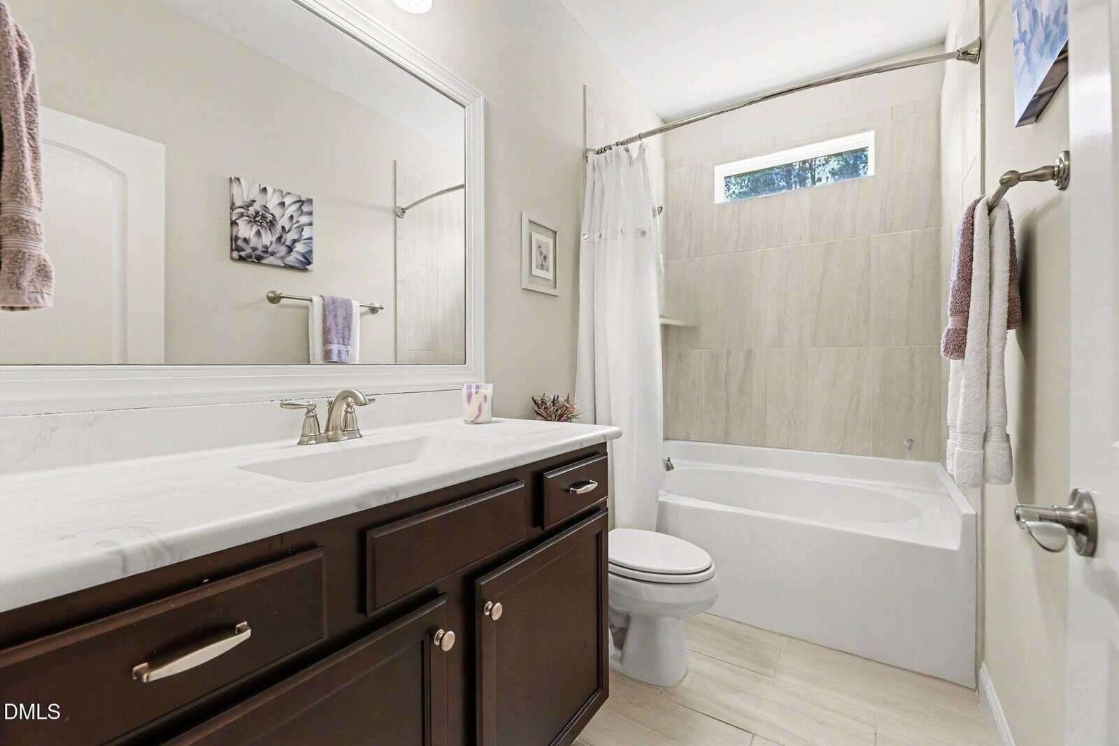 Modern bathroom with dark wood vanity, white countertop, large mirror, bathtub with beige tiles, white shower curtain, and two towels on racks. Natural light comes through a high window above the tub.