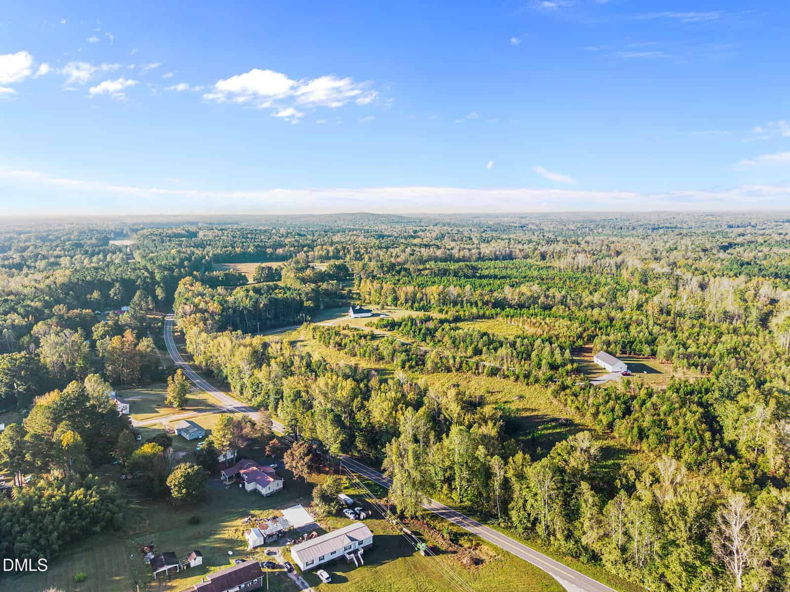 Aerial view of a rural landscape with scattered houses, winding roads, dense green trees, and open fields under a clear blue sky.