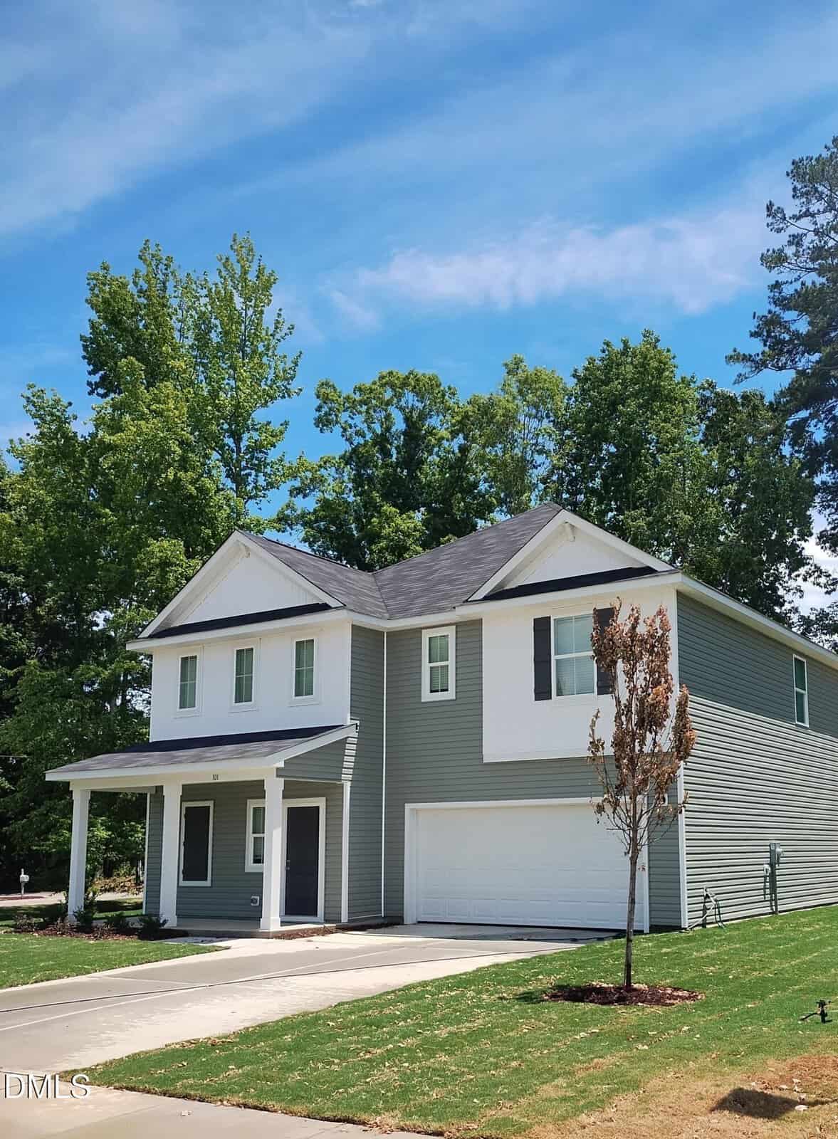 Two-story modern house with gray siding, white trim, and a two-car garage. The home has a front porch, a concrete driveway, and is surrounded by green trees under a blue sky with wispy clouds.