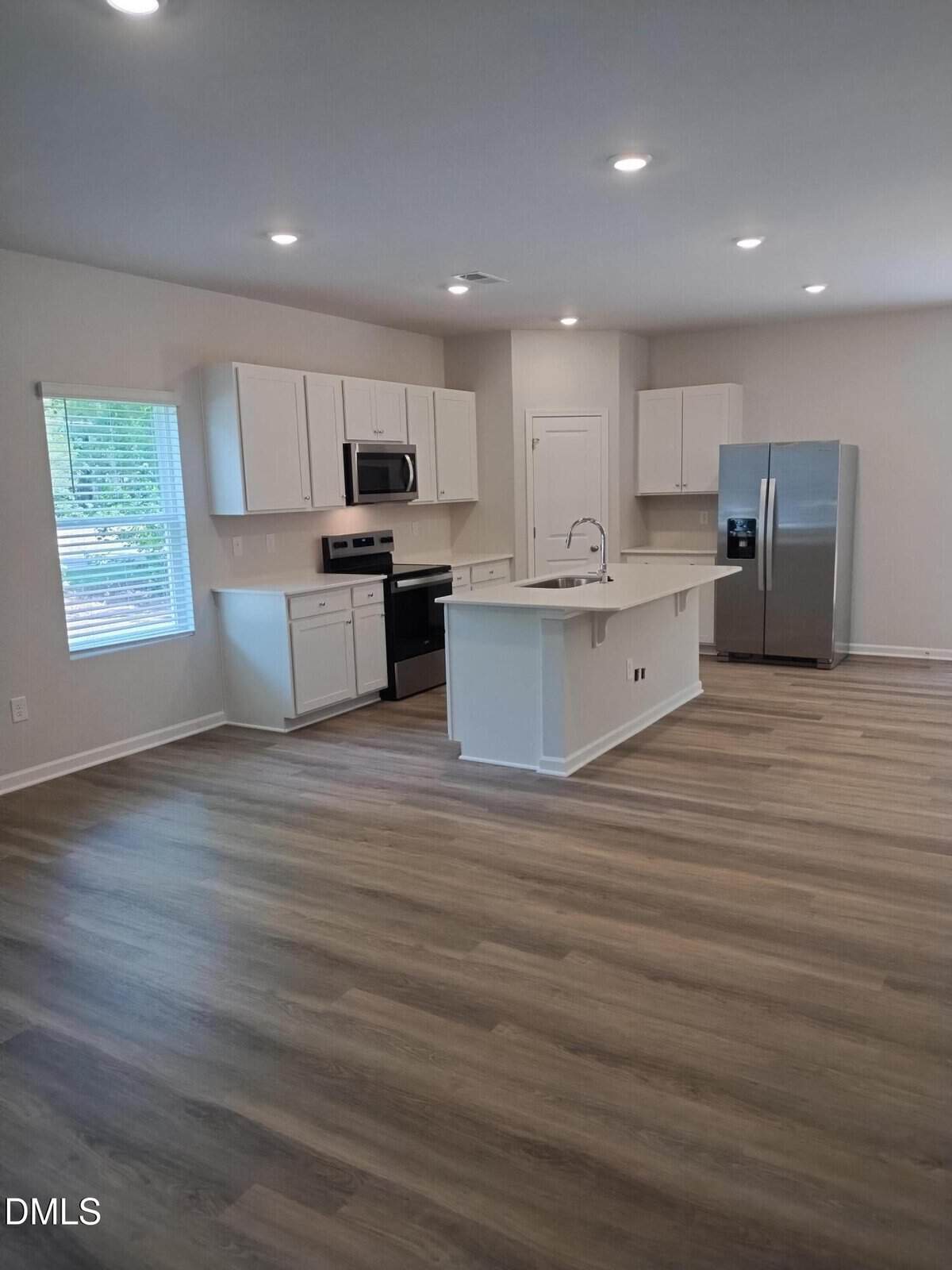 Modern kitchen with white cabinets, stainless steel appliances, a center island with a sink, light wood flooring, and a window letting in natural light. Walls are painted a light neutral color.