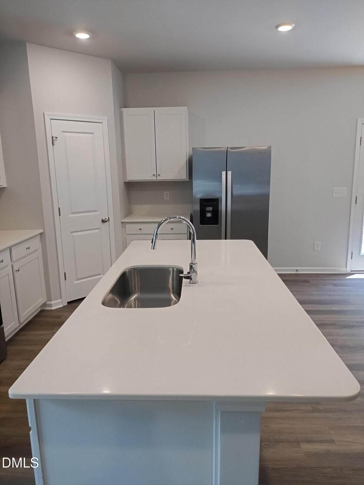 Modern kitchen with a large white island featuring a built-in sink and faucet, stainless steel refrigerator, white cabinets, and wood-style flooring under bright ceiling lights.