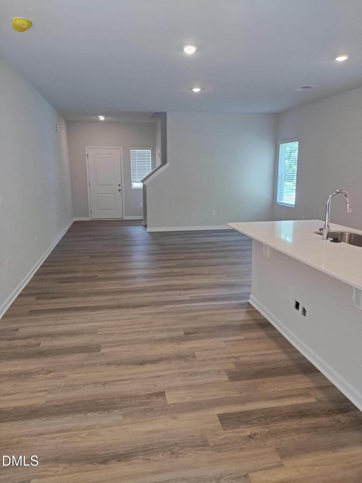 Modern, unfurnished open-plan living space with wood flooring, white walls, recessed ceiling lights, and a kitchen island with a sink; a door and window are visible in the background.